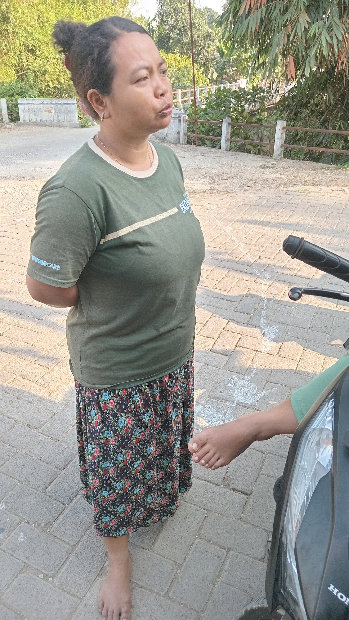 In Kedungwaru, Indonesia, a woman with dark, curly hair pulled back stands on a paved walkway. She is wearing a green t-shirt with a logo on the sleeve and a floral-patterned skirt. Her arms are crossed behind her back. To her right, a person's bare foot is visible, resting near a motorcycle. The environment appears to be outdoors, with trees and foliage in the background.