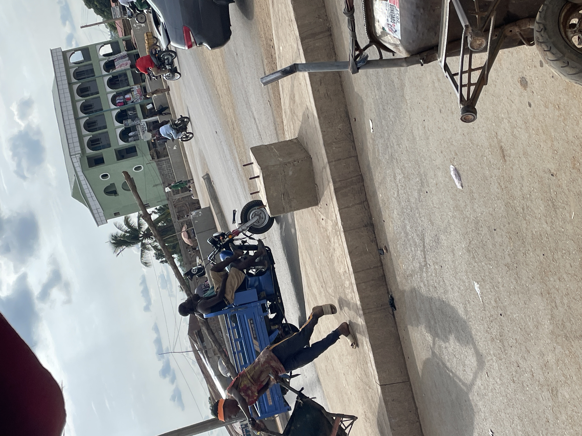 The image depicts a street scene in Akwanga, Nigeria. A light green multi-story building with arched windows and decorative circles is visible in the background, situated on the left side of the frame. Several people on motorbikes are also visible, one with a red shirt. In the foreground, a person pulls a small, green cart along the side of the road. Another person rides on a blue three-wheeled motorcycle, a common mode of transport in the area. A cement curb is visible. In the upper right corner, the tire of a vehicle is seen, along with a sign labeled "Governor 2023". The scene seems to be taken during the daytime, under a partly cloudy sky.