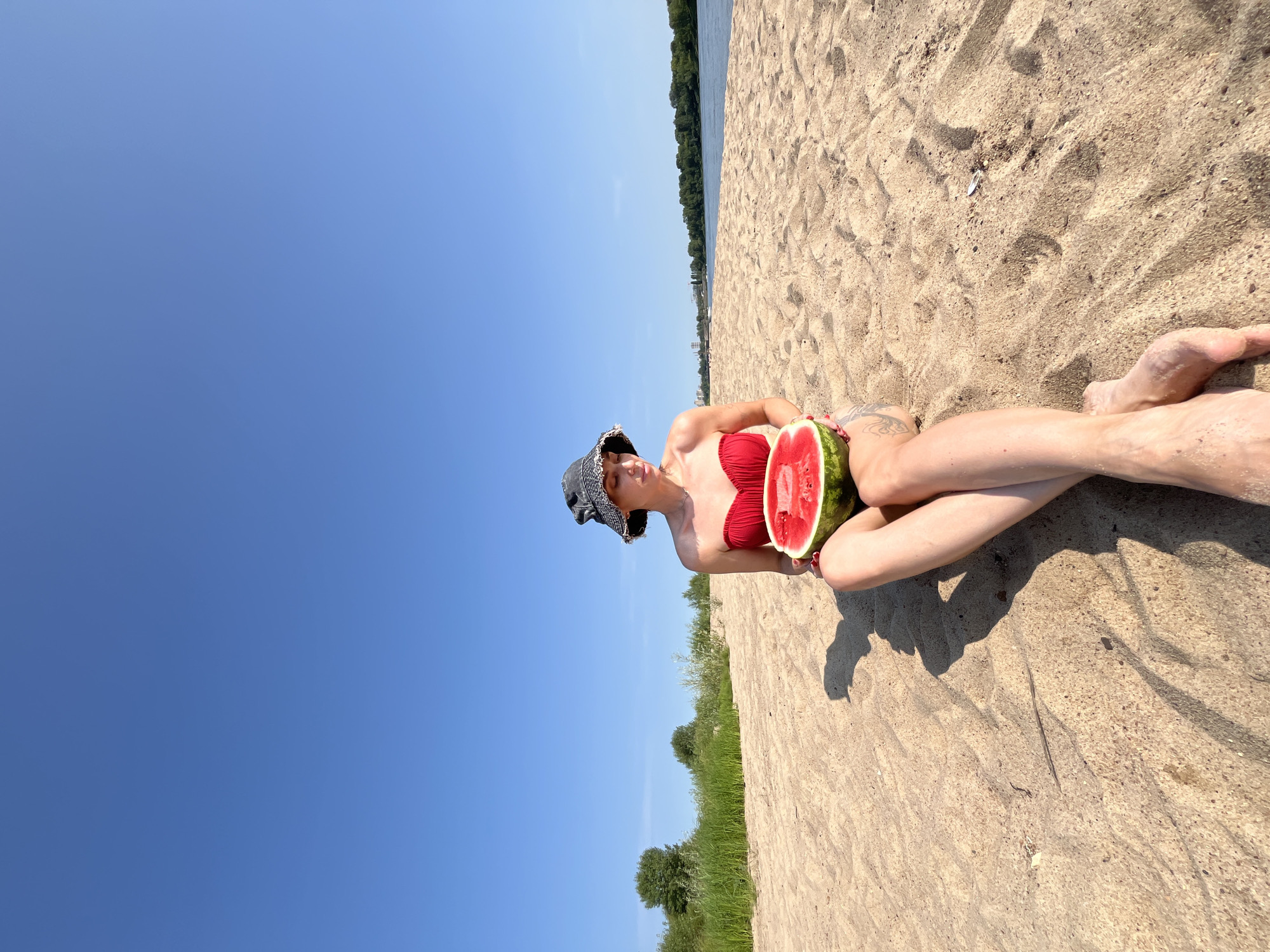 A woman wearing a red bikini top and a bucket hat sits on a sandy beach with a half-eaten watermelon in her lap. The woman is looking down at the watermelon. The background is a clear blue sky and a body of water with trees on the opposite bank. The scene appears to be the beach on the Volga River in Nizhny Novgorod, Russia. The sand is light brown and textured with small pebbles and debris. The lighting suggests it is daytime, and the sun is casting shadows on the sand.