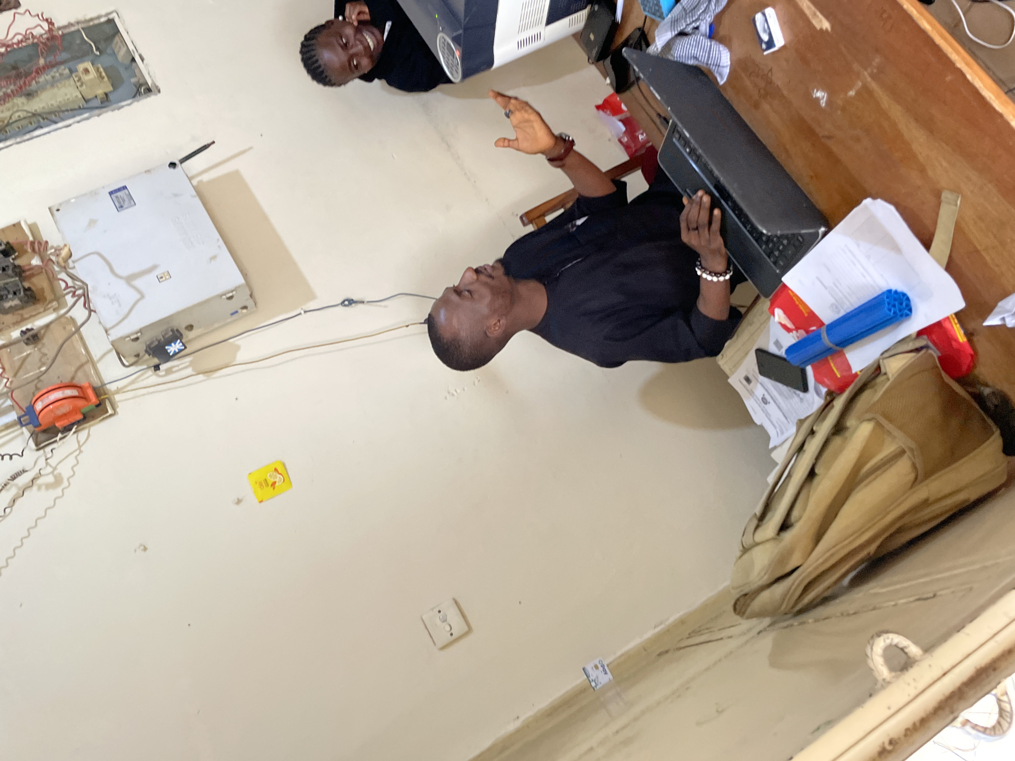 In Durumi, Nigeria, a man wearing a black traditional outfit sits at a wooden desk, engaging with a laptop and gesturing with his right hand. To his right, a woman in a black hooded sweatshirt looks at a computer monitor and smiles. The desk is cluttered with various items, including papers, a blue cylindrical object, a mobile phone, and a beige backpack leaning against the wall on the left. The background features a beige wall with electrical conduits, circuit breakers, and a yellow sticker with a drawing. The scene appears to be an indoor office or workspace.