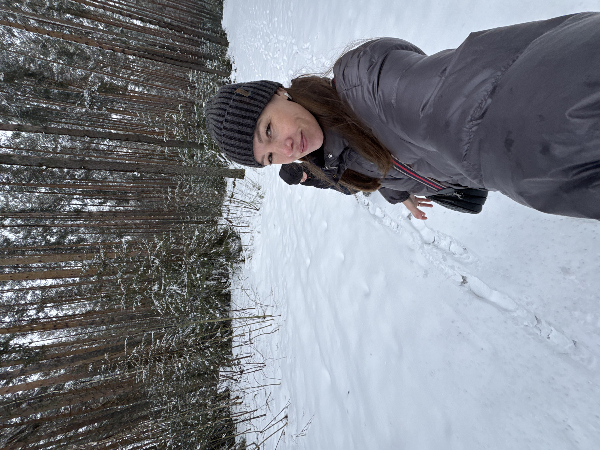 A woman in a gray jacket and striped beanie stands in a snowy environment next to a dense forest. The location is Chaikovsky, Russia. The woman appears to be taking a selfie. Her left hand is gloved. Footprints are visible in the snow.