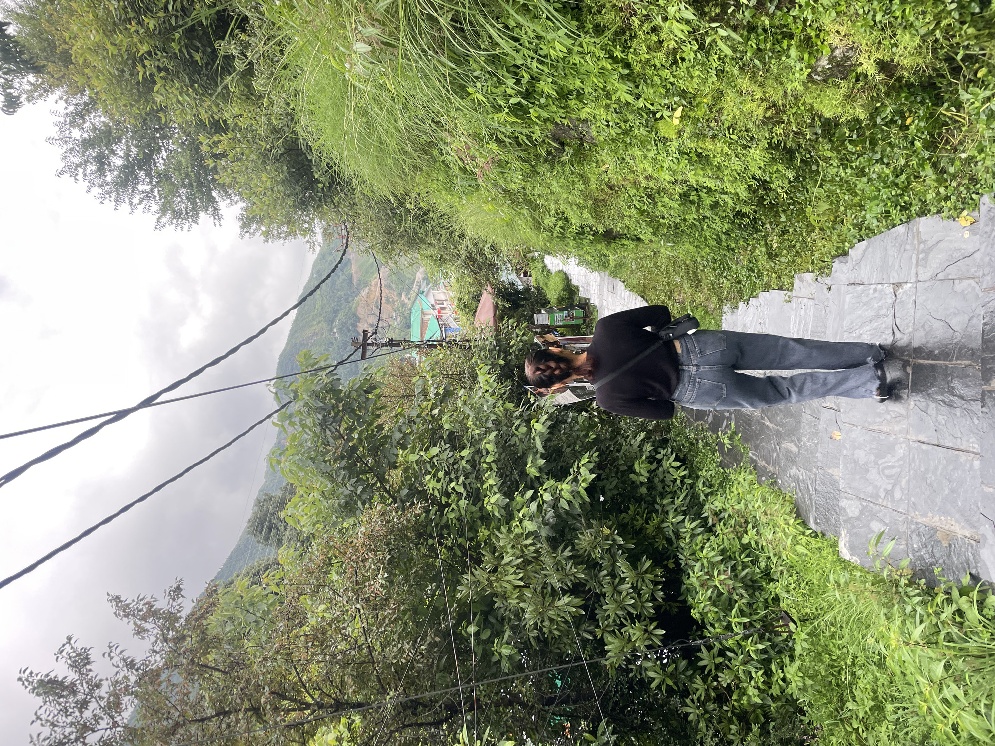 A person wearing a black top and blue jeans walks down a wet, stone staircase in Bhagsu, India. The staircase is lined with lush green vegetation on the right and trees on the left. In the background, there are buildings with colorful roofs and a mountainous landscape under a cloudy sky. Overhead, power lines are visible.