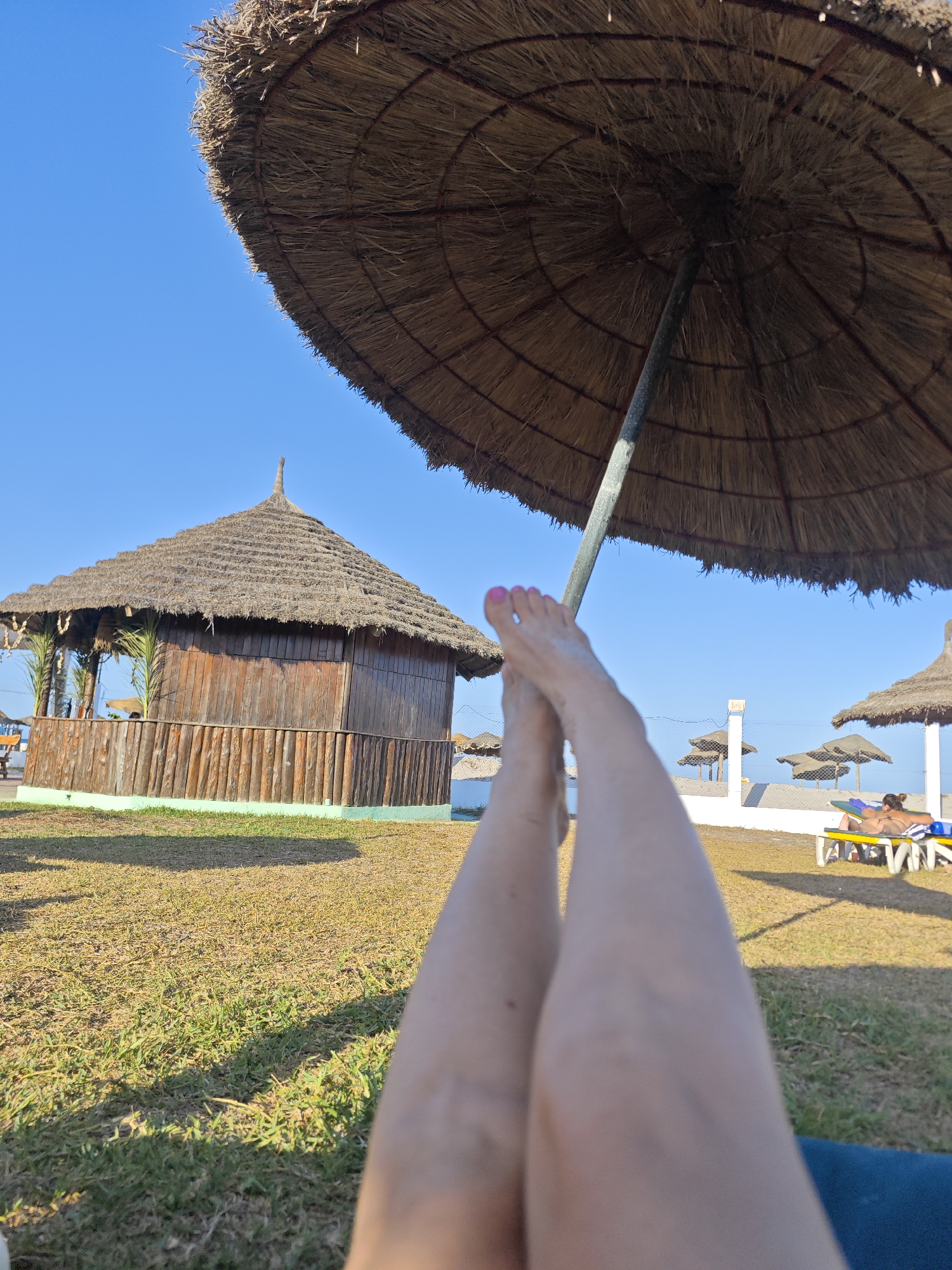 The image depicts a beach scene in Hiboun, Tunisia, under a clear blue sky. In the foreground, a person's legs are crossed, resting on a surface that suggests a beach chair. A large, straw umbrella provides shade from the sun, its textured underside and central pole visible.
In the middle ground, a rustic wooden hut with a thatched roof stands on a grassy area. The hut has a fence made of vertical wooden logs around its base. Palm fronds are visible behind the hut. Several other straw umbrellas dot the landscape, some casting shadows on the grass.
In the background, a person is seen relaxing on a yellow and blue lounge chair under one of the umbrellas. A white fence runs horizontally across the mid-background. The overall setting is a beach resort or a similar recreational area, characterized by the natural elements of grass, sand, and sun, along with man-made structures for shade and relaxation.
