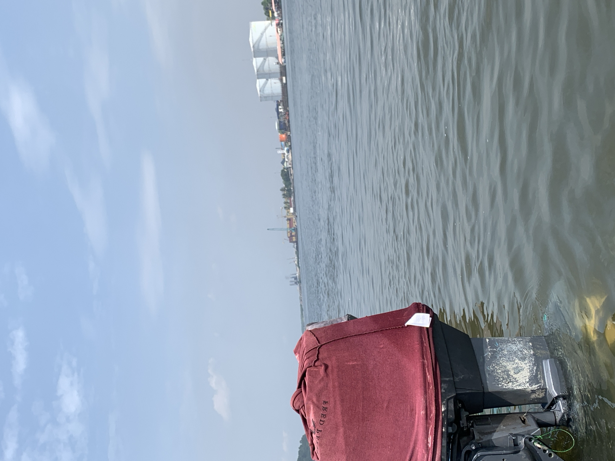 The image shows a view from a boat on the water in Ibasa, Nigeria. The water is gray and choppy. In the background is a shoreline with buildings and industrial structures, including large white storage tanks. The sky is light blue with sparse clouds. In the foreground, part of the boat's motor is visible, covered by a burgundy fabric.