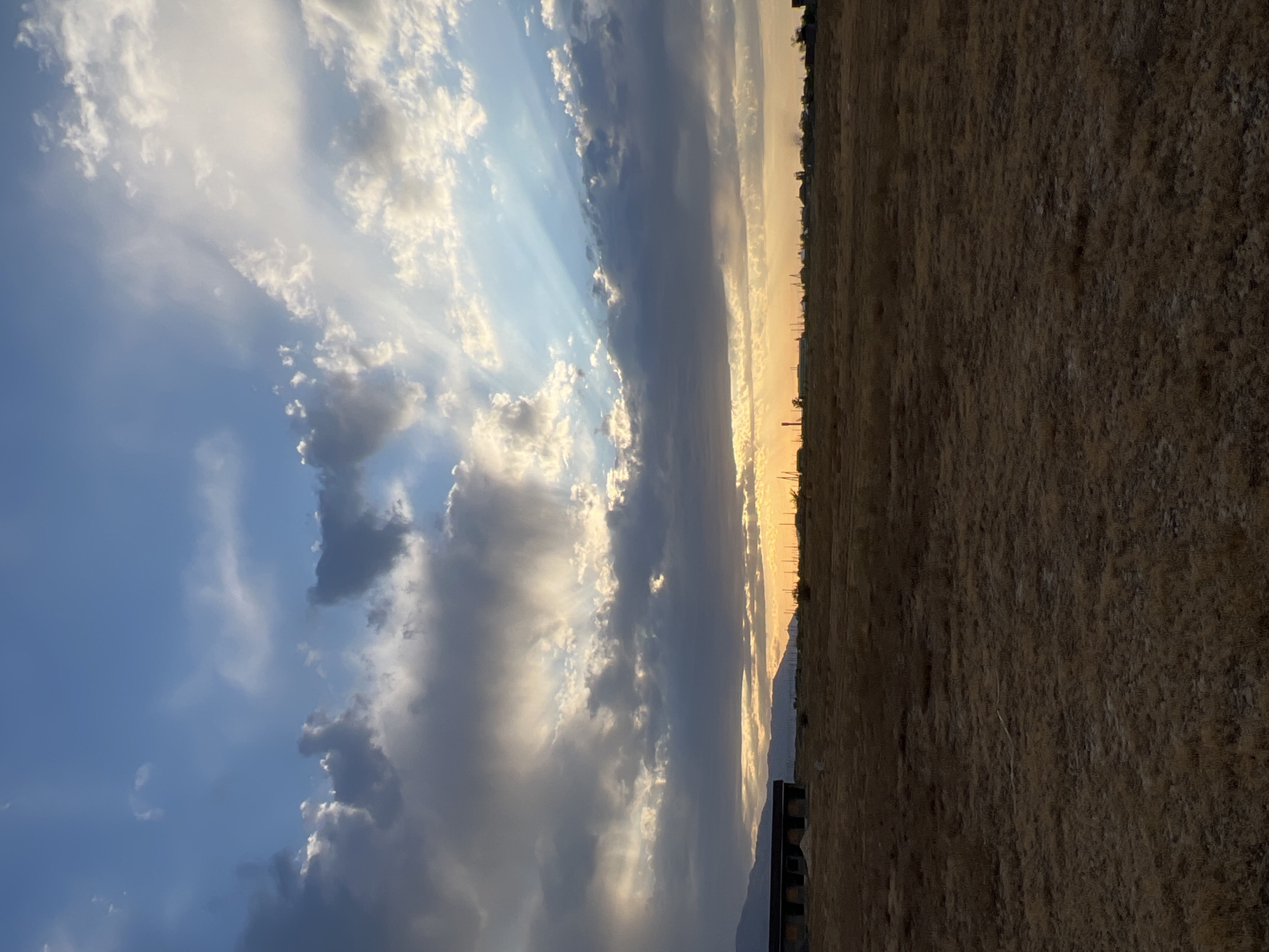 The image shows a wide, flat, dry, grassy field under a dramatic sky. Dark, heavy clouds are interspersed with patches of bright blue sky and shafts of sunlight breaking through. The sun is low on the horizon, casting a warm glow that illuminates the edges of the clouds and creates a golden band across the distance. In the far distance, a low, dark silhouette of a town or settlement is visible, with some structures and possibly trees. To the left, a partially constructed, dark-colored building with visible arched openings is situated closer to the foreground. The ground is covered in dry, light brown vegetation, appearing parched. The overall scene suggests an arid or semi-arid environment, possibly at sunset or sunrise. The location is identified as Ақши, Kazakhstan. There are no people or animals visible in the image.