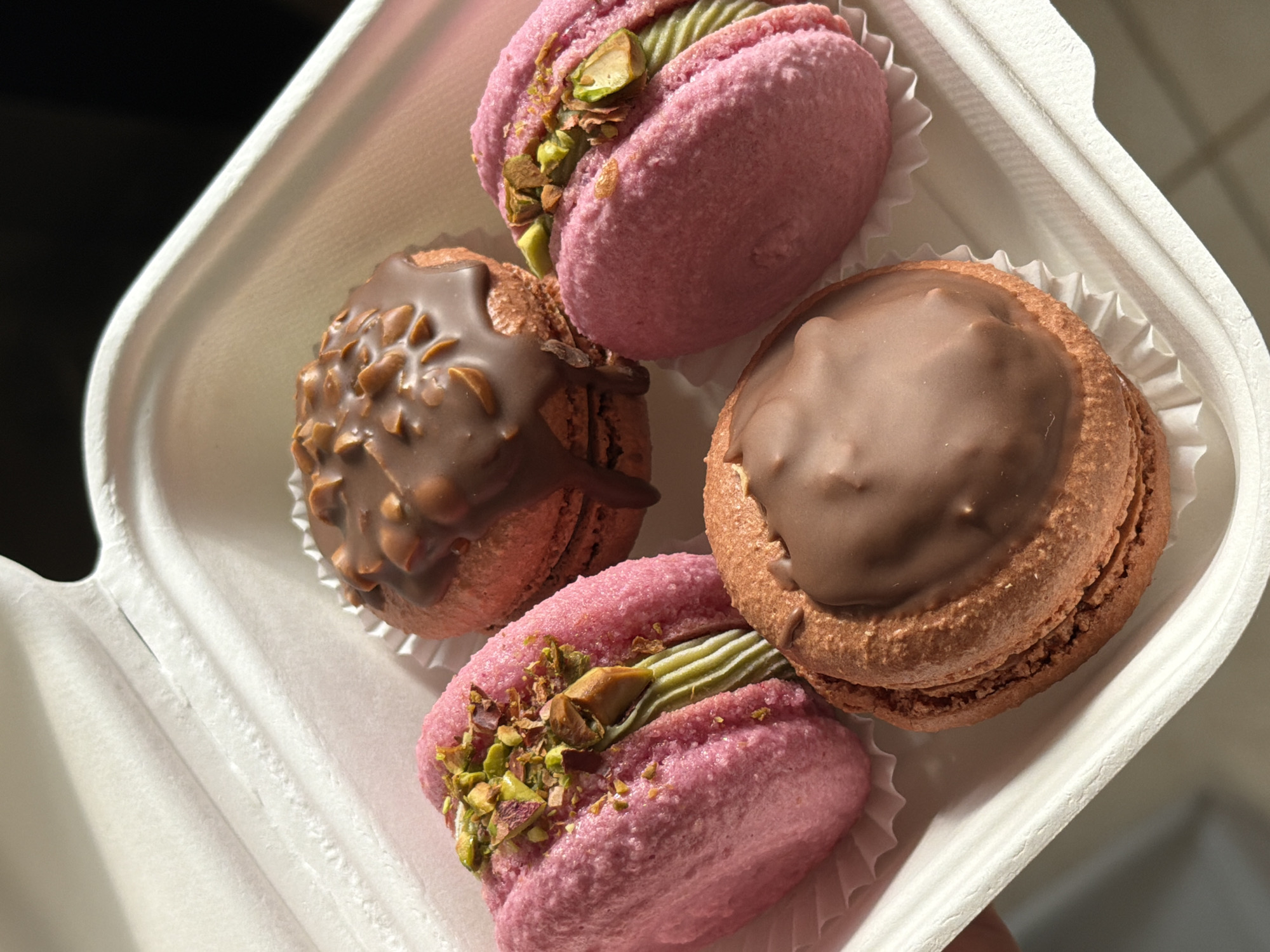The image depicts a white container holding four macarons. Two of the macarons are pink and feature a green filling topped with chopped pistachios. One macaron is brown with a smooth chocolate ganache on top, while the other is brown with a chocolate ganache decorated with small pieces of nuts. All macarons are in white paper cups within the container. The photo appears to be taken indoors with natural light. There is no information to suggest that this image was taken in Tel-Aviv, Israel.