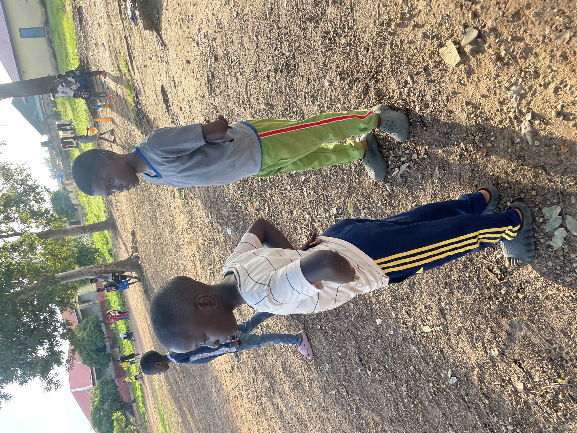 The image depicts an outdoor scene with several young boys in the foreground and background. The ground is predominantly dirt and gravel, with sparse vegetation. In the foreground, two boys are positioned to the left and center. The boy on the left is partially visible, wearing a blue and white striped shirt and dark pants. The boy in the center, facing right, is wearing a light-colored, striped t-shirt with dark blue pants adorned with yellow stripes. He has his hands on his hips. To the right of the center boy, another boy is standing, facing left, wearing a grey t-shirt and bright green pants with a red stripe. He is wearing dark clogs. In the background, several other children are visible, some interacting and others standing. Behind them, there are trees, and in the distance, buildings with red and yellow roofs are present. The lighting suggests it is daytime, with shadows cast on the ground. The environment appears to be a somewhat informal outdoor area, possibly a schoolyard or a community space, in Jos, Nigeria.