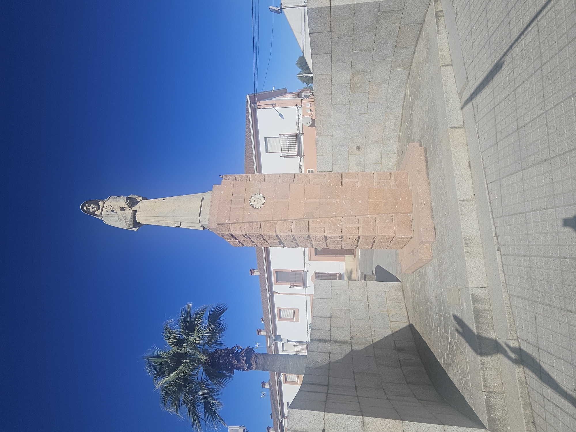 This image depicts the statue of Abad Pedro. The statue stands on a tall, reddish-brown rectangular base with a round medallion on its side. The base rests on a gray stone platform in the city of Pedro Abad, Spain. A palm tree grows nearby. The sky is clear and blue. Buildings are visible behind the monument. The scene is captured in bright daylight. A shadow of the statue is cast on the ground.