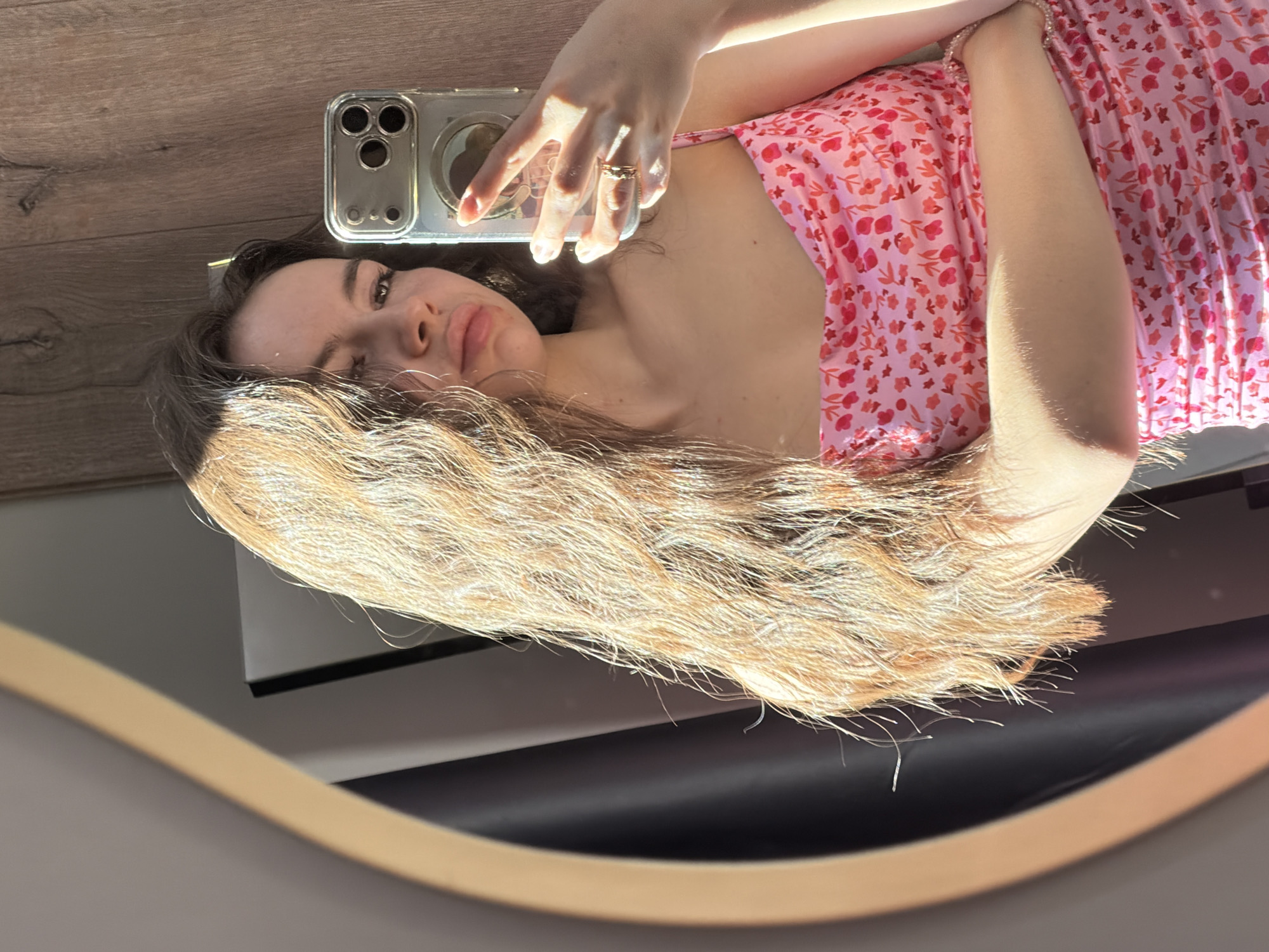 A young woman takes a selfie in a mirror. She is wearing a pink dress with a floral pattern. She holds a silver smartphone in her right hand. Her long brown hair is partially obscuring her face. The background includes a wooden floor and a black surface. The location cannot be determined from the image.