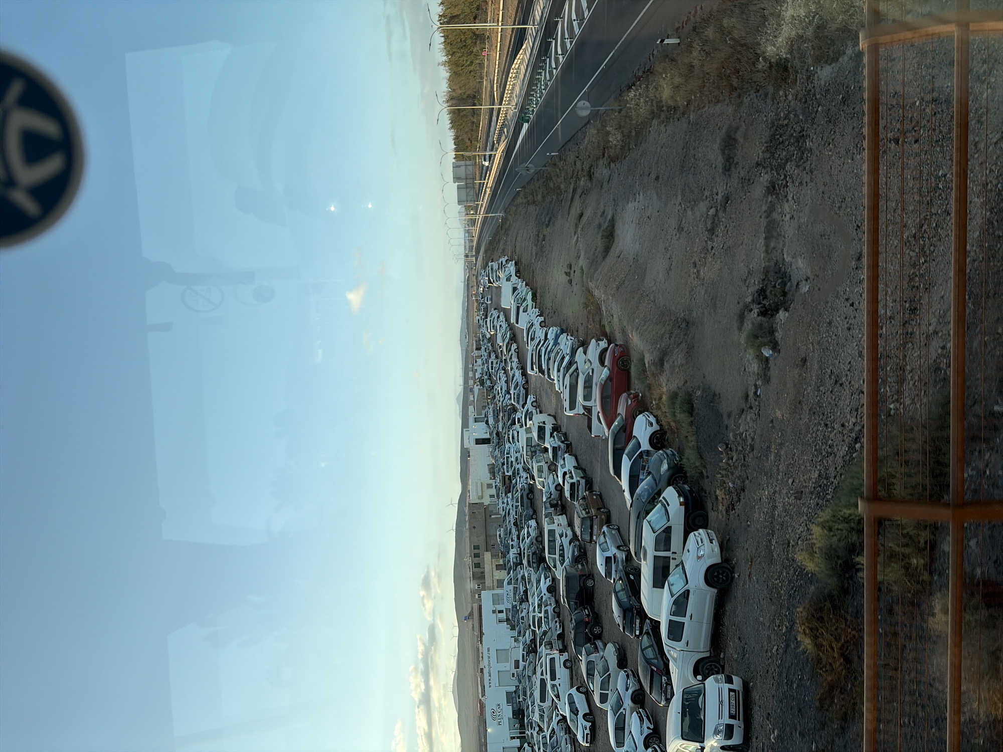 The image shows a large parking lot filled with primarily white vans, located next to a highway. The vans are parked in rows on a dirt surface. In the background are buildings and hills, potentially indicating an industrial or commercial area. The overall scene is shot from an elevated perspective, possibly from a building window, with part of the window frame visible. Given the clues present such as the website on the side of one of the vans, it is likely the image shows the industrial estate of Puerto del Rosario, Spain.