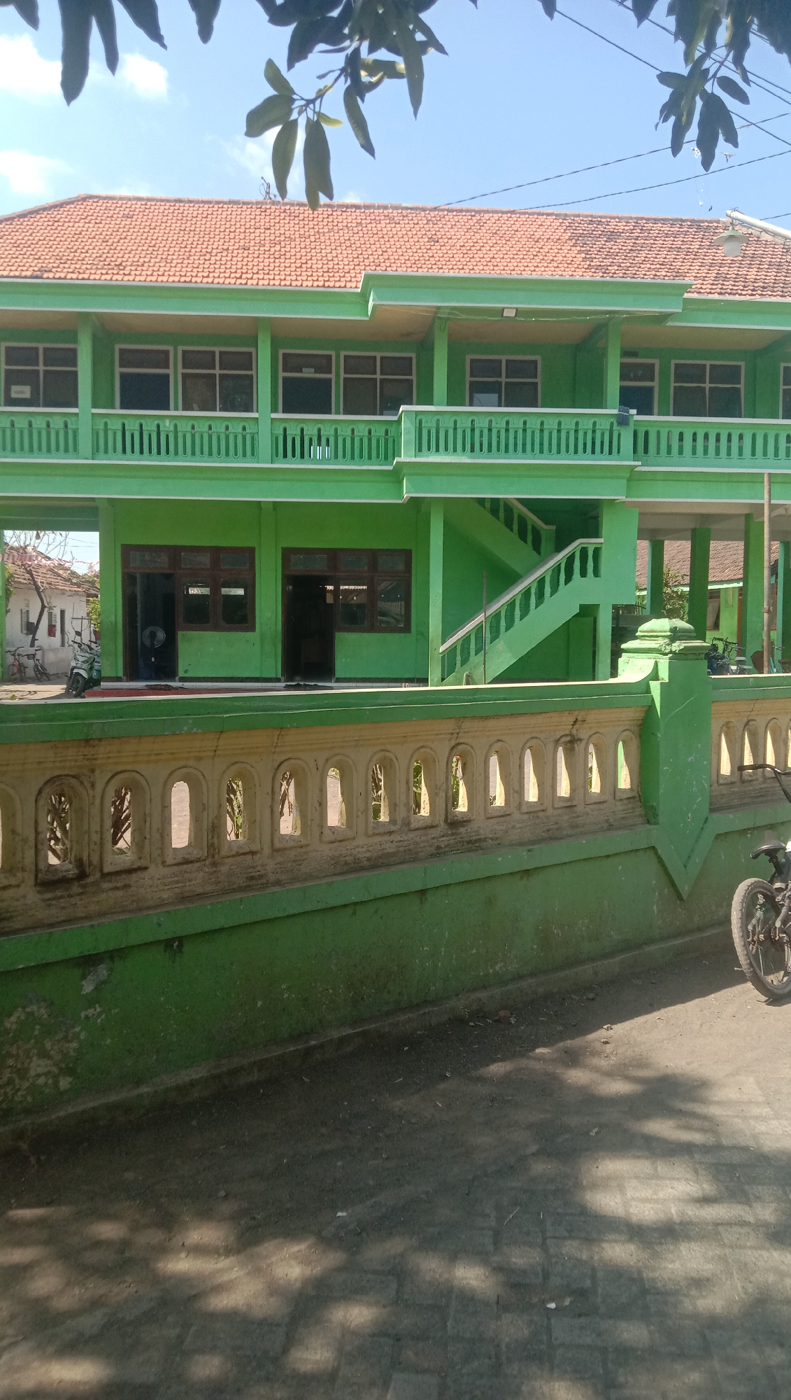 The image shows a two-story green building with a red tile roof, located in Ngempit, Indonesia. The building has a balcony on the second floor with a green railing and a decorative white fence in front of it. There are several windows on both floors, and two open doorways on the ground floor. A staircase leads to the second floor. In front of the building, there is a paved area with some shadows cast by trees. A motorcycle is parked to the right of the image, partially visible. The overall scene depicts a school or an institutional building under a clear blue sky.