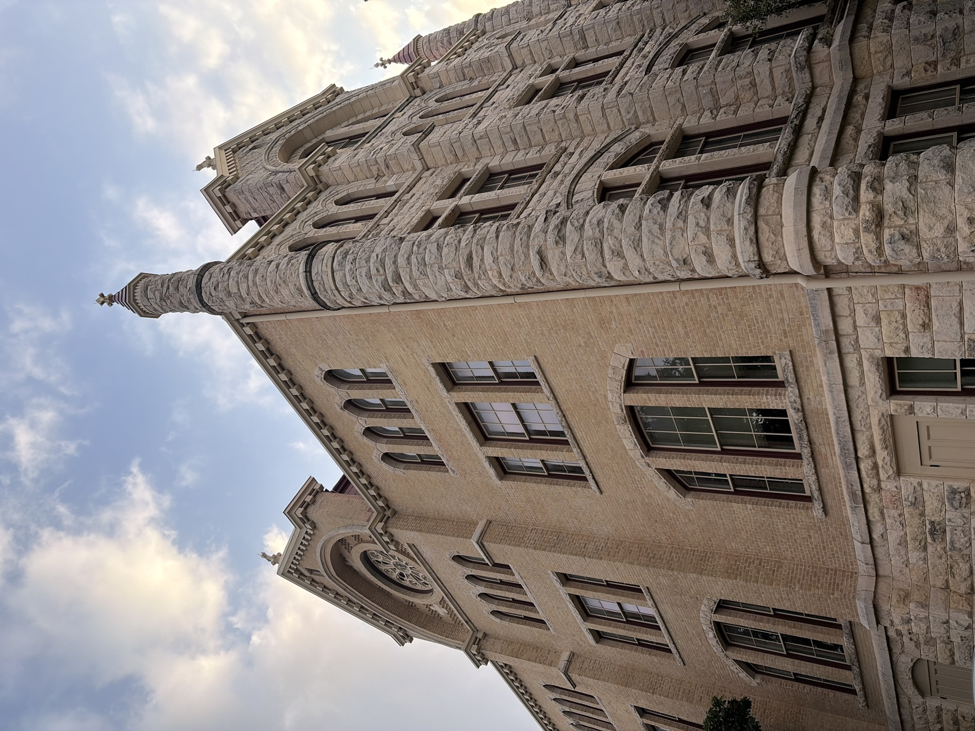 The image shows a low-angle view of the Texas State Capitol building in Austin, Texas, United States. The building's facade is prominently featured, showcasing its architectural details, including arched windows, stone embellishments, and a decorative tower. The sky with some clouds provides the backdrop.