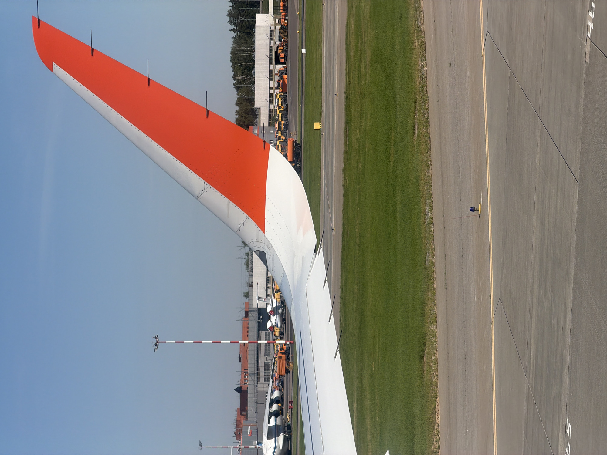 The image shows a view from an airplane window at an airport, likely Sheremetyevo International Airport in Khimki, Russia, based on its characteristic tail fin design. The primary focus is on the wing of the airplane, which features a white underside and a striking orange and white vertical stabilizer. In the background, the airport tarmac is visible, with several parked aircraft, including a large cargo plane and smaller passenger jets. Various airport service vehicles, such as orange trucks and yellow construction equipment, are also present. Airport infrastructure, including buildings, hangars, and tall striped light poles, are scattered across the scene. The environment appears to be a sunny day with a clear blue sky. There are no people clearly visible in the image.