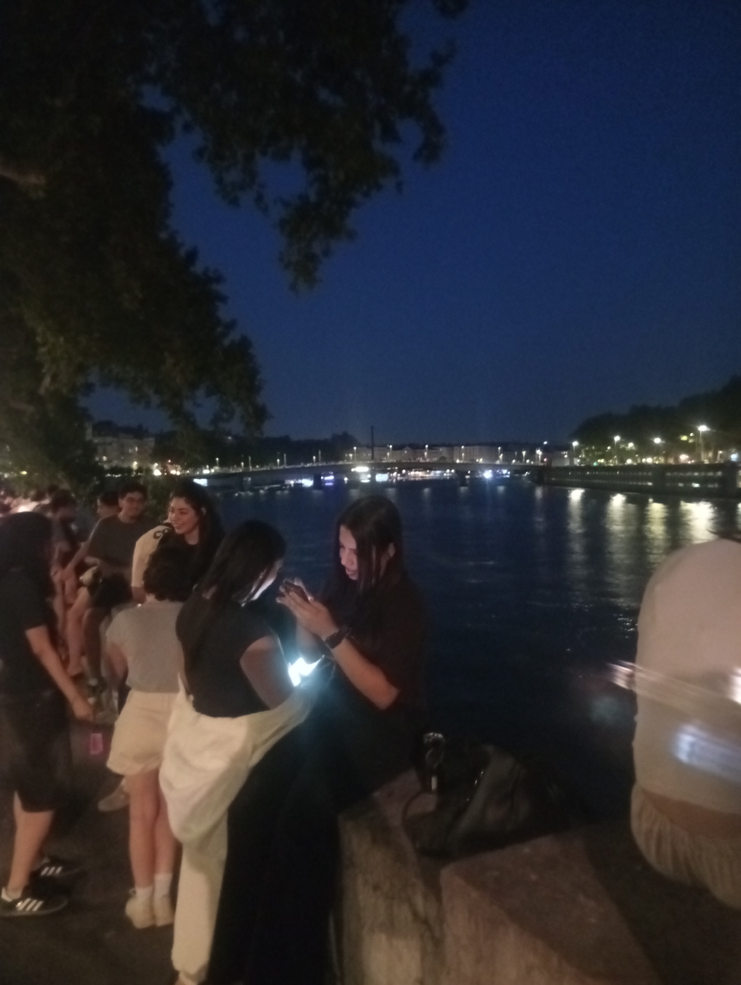 The image captures a nighttime scene along the Saône River in Lyon, France, possibly during a festival or event. A group of people are gathered on a stone embankment, illuminated by streetlights and the glow of their mobile phones. The water of the river reflects the city lights, creating a shimmering effect. In the background, buildings and bridges line the opposite bank, with more lights visible in the distance. The atmosphere appears relaxed and social, with individuals engaged in conversation and using their devices. The presence of trees and the dark sky suggest it is late evening or early night.