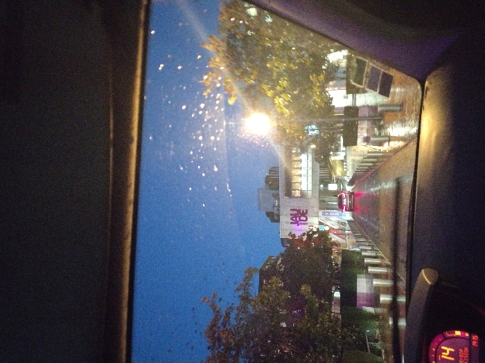 The image presents an urban evening scene viewed from the interior of a vehicle, looking forward through a rain-splattered windshield. The vehicle's dashboard, visible in the lower foreground, features a digital display showing "OCTOBRE" and "19:15" (or "19:16"), indicating the time and month. The central focus is a multi-story building displaying the prominent, illuminated sign "JAUDE" in large letters, identifying it as Centre Jaude in Clermont-Ferrand, France. A wet, reflective paved path, bordered by numerous silver cylindrical bollards, leads directly towards the building's entrance. A dark-colored sedan with illuminated red taillights is positioned centrally on this path, facing away from the viewer. The environment is illuminated by streetlights, which cast reflections on the wet ground, and the building itself is brightly lit. Trees with a mix of green and autumnal yellow foliage are visible on either side of the path. The sky is a deep blue, characteristic of dusk or night. No people are visible.