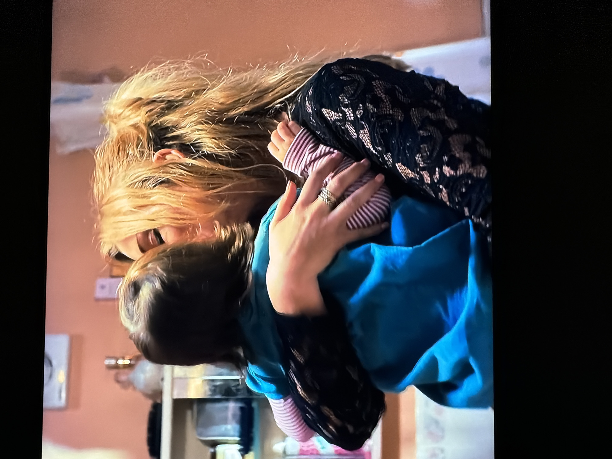 The image captures a tender indoor moment between a woman and a young child, set in what appears to be a home in Sofiivska Borshchahivka, Ukraine.
The woman, with long, light blonde hair that appears slightly tousled, is seen from a side-rear angle, embracing the child. She is wearing a black top or dress with a distinct lace pattern. A prominent ring is visible on her left ring finger. Her face is partially visible, showing one eye and parts of her nose and mouth, suggesting a gentle expression as she looks towards the child.
The child, likely a baby or toddler, has dark hair and is held close to the woman's chest and shoulder. The child wears a light-colored, long-sleeved top with thin horizontal stripes, possibly pink and white or red and white, layered with a bright blue garment, possibly a jacket, blanket, or carrier.
The activity depicts a close embrace, conveying a sense of warmth and affection between the two individuals.
The setting is an indoor space with a warm, light brown or peach-colored wall. In the background, on the right side, a stainless steel object, possibly part of a sink or kitchen appliance, is visible. On the left side of the frame, a blurred white rectangular object, possibly a wall-mounted thermostat or light switch, can be seen. The lighting suggests an ordinary indoor environment, not indicating a specific time of day. There is no visible text in the image.
