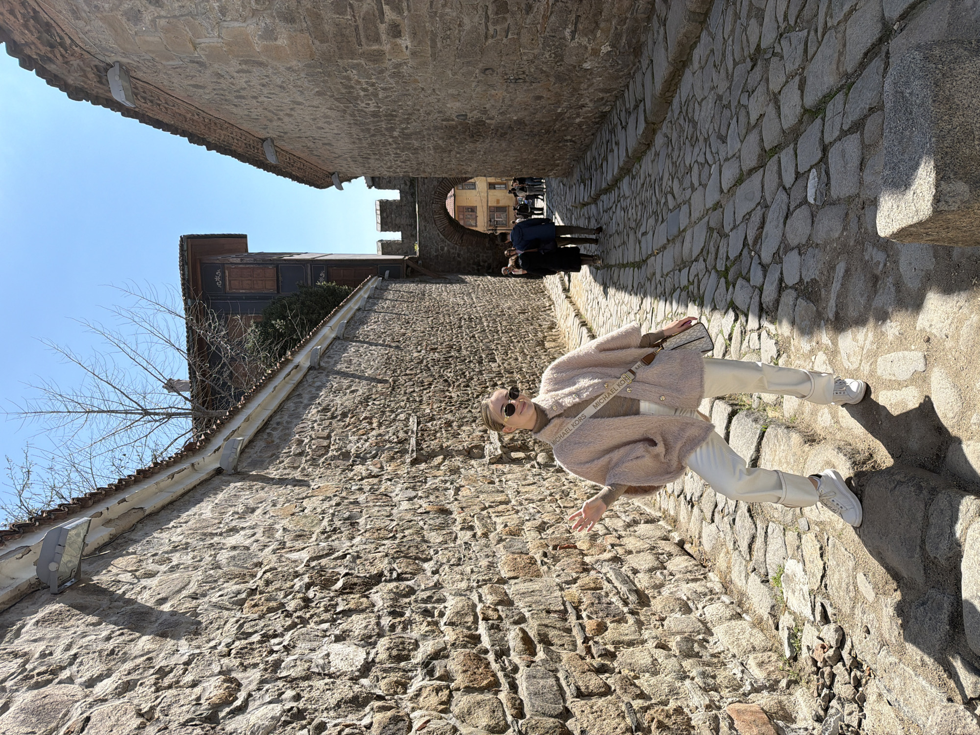 A woman is standing in a narrow, cobbled street in Plovdiv, Bulgaria. She is wearing a light pink jacket, white pants, and white sneakers, and she has a crossbody bag. She is also wearing sunglasses and has her arms outstretched. The street is lined with old stone buildings. A group of people is seen in the background walking through a stone archway. The sky is bright blue.