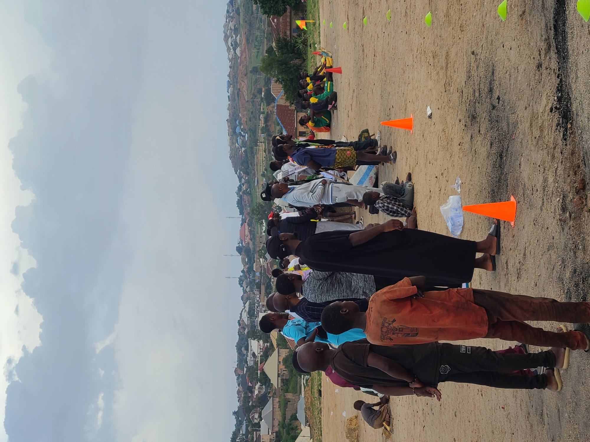 The image shows a group of people, predominantly young men and boys, gathered on a dirt field. They are lined up, some standing and some kneeling, facing away from the camera. The ground is sandy with some scattered debris. Orange traffic cones are visible in the foreground and scattered further back, suggesting a sporting event or training session. In the background, there are houses and buildings on a hillside, characteristic of an urban or semi-urban environment in Jos, Nigeria. The sky is overcast, indicating cloudy weather. The people are dressed casually, in t-shirts, shorts, and trousers. Some are looking upwards, possibly at an instructor or during a waiting period. The overall impression is of an organized outdoor activity. No text is clearly visible on any of the people or objects. The time of day appears to be daytime, given the general illumination.