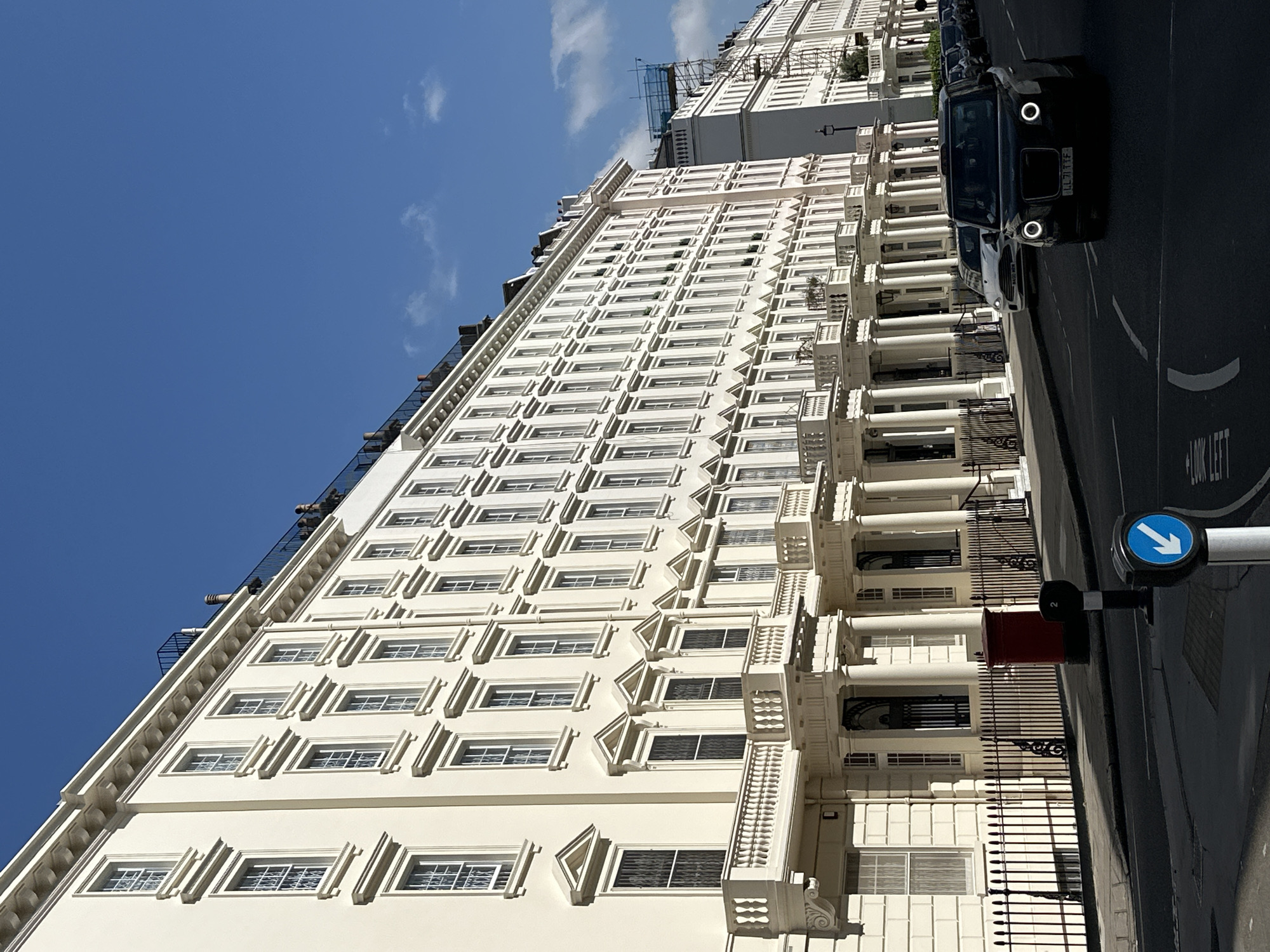 The image shows a street scene in London, United Kingdom, featuring a long, multi-story white building with many windows arranged in rows. The building's architecture includes decorative elements around the windows and classical-style columns supporting small balconies. A black London taxi is parked on the street alongside the building. A street sign with a blue arrow indicating the direction of traffic is also visible. The sky is clear and blue.
