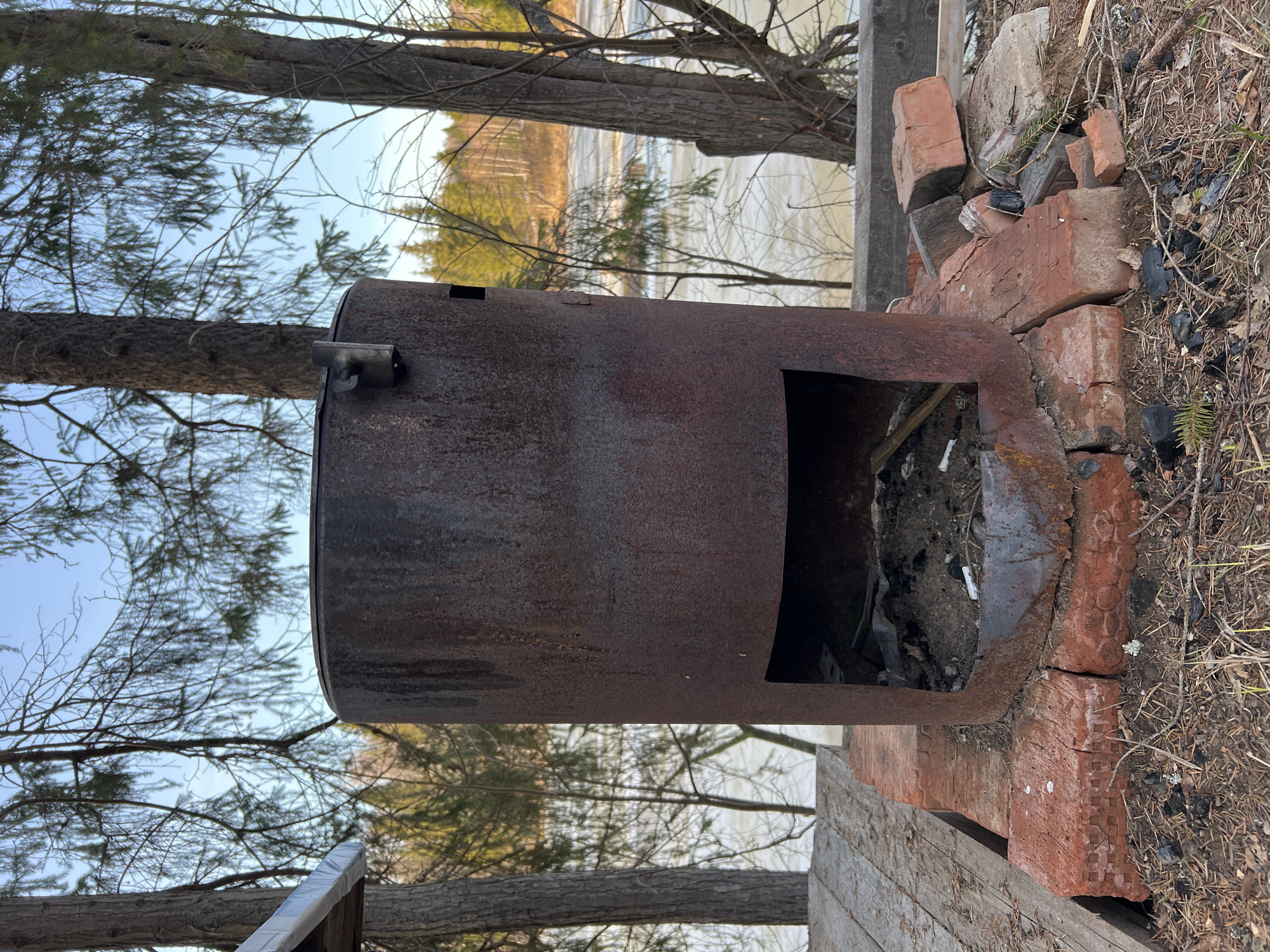 A rusty, cylindrical metal incinerator sits outdoors on a base of red bricks. The incinerator has a square opening near the bottom and a small protrusion at the top. Debris is visible inside the incinerator. The bricks are arranged in a disorganized manner. The surrounding environment includes trees, dry grass, and a body of water with vegetation visible in the distance. The location is Zhigalki, Russia.