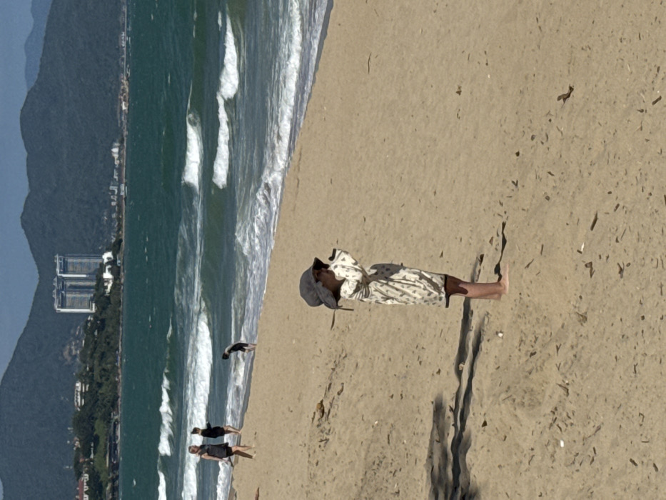A high-angle shot shows a beach with a person wearing a light-colored patterned outfit and a hat lying horizontally. Their head is towards the left, near the ocean. Two other individuals and a bird are near the water's edge. Waves are visible in the ocean, and there's a mountain range and buildings in the distance. The sand is a light color and displays various debris and shadows.