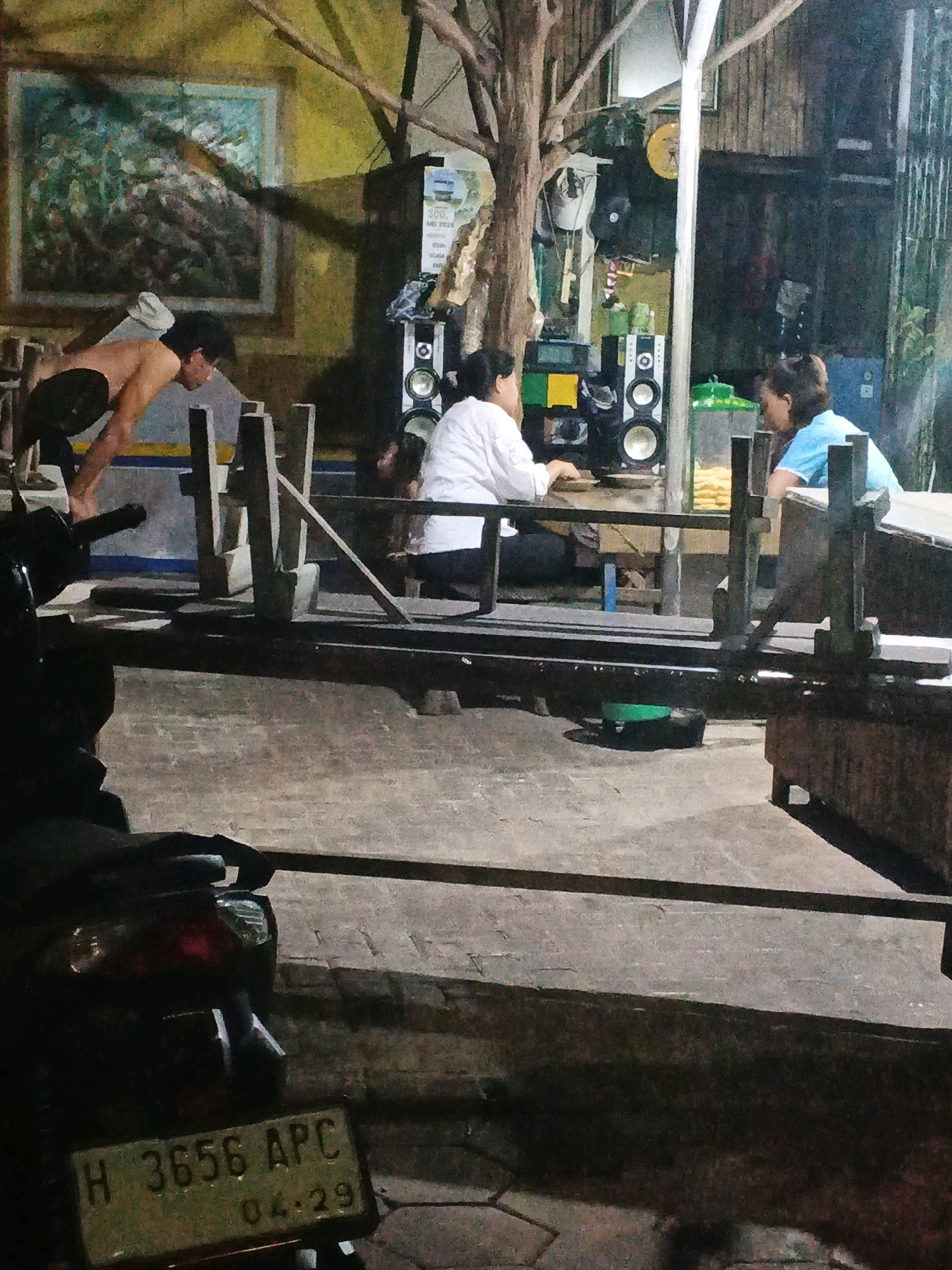 The image depicts an outdoor scene at night, possibly in Ungaran, Indonesia, characterized by dim lighting and a generally unkempt environment. In the foreground, the rear of a dark-colored motorcycle is visible, featuring a red taillight and a green license plate displaying "H 3656 APC" and "04.29". A group of people are gathered in the middle ground. On the left, a shirtless individual is positioned in a squatting or bending posture. Further back, two individuals are seated at a table or counter. One person, wearing a white garment, is facing away from the camera, while another, in a light blue shirt, is seen from the side. Behind them, a sound system with two speakers is set up. The background shows wooden structures and some greenery. The ground appears to be paved with interlocking bricks. The overall impression is of a casual, possibly informal gathering in an outdoor, dimly lit setting.