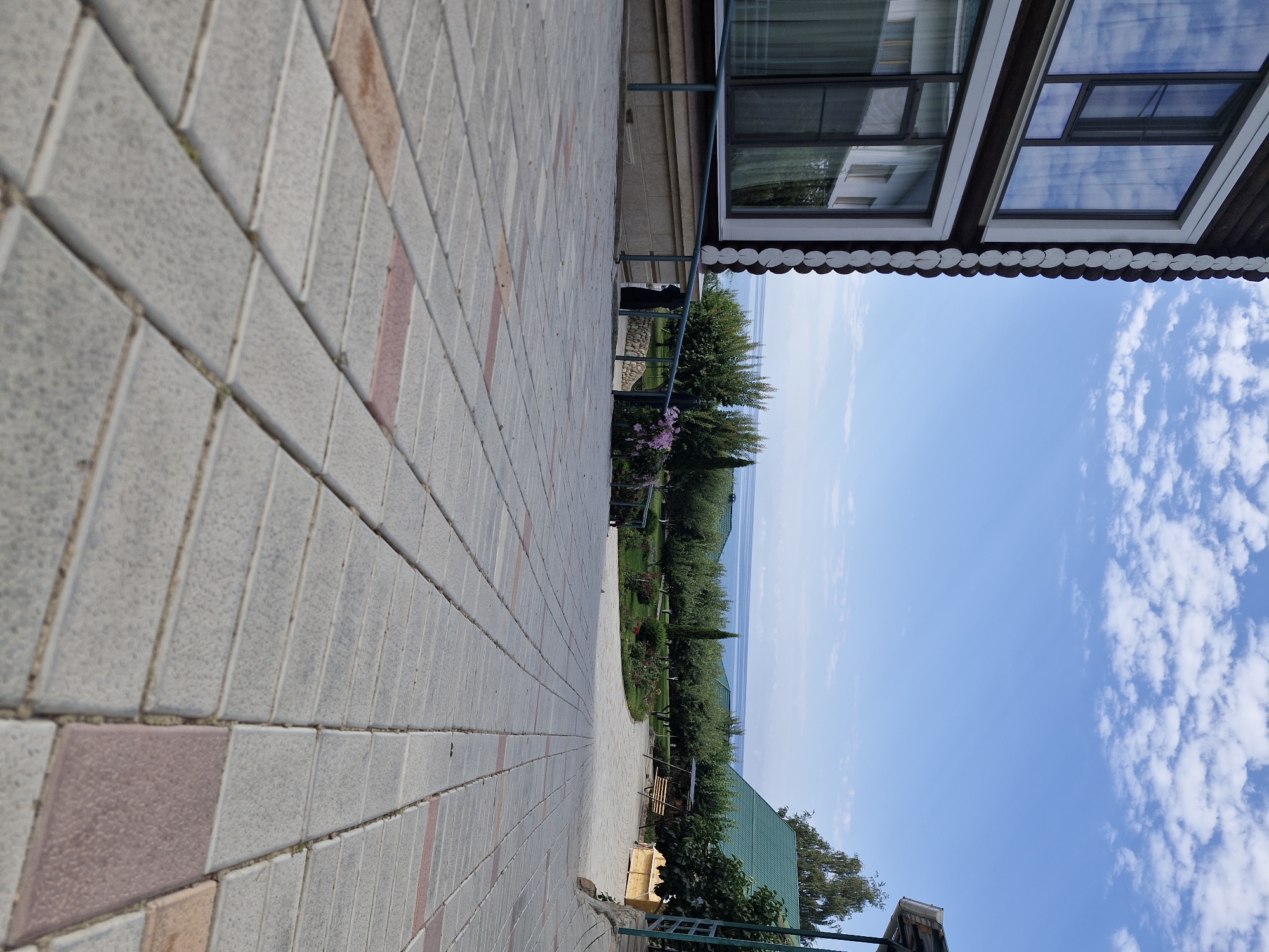 The image depicts a paved walkway leading towards a body of water, likely Issyk-Kul Lake, given the context of Bulan-Segettu, Kyrgyzstan. A multi-story wooden building with dark brown siding and white trim is visible on the left, featuring large windows reflecting the sky and clouds. A metal railing runs along a ramp beside the building. In the background, a well-maintained garden with trees, bushes, and flowers leads to the lake. A green-roofed building and a wooden swing set are also present in the garden area. The sky is a vibrant blue with scattered white clouds. The walkway is constructed of rectangular grey and red-brown paving stones, laid in a staggered pattern. The low-angle perspective emphasizes the texture and pattern of the paving stones. The scene suggests a peaceful, resort-like environment. There are no people visible in the image.
