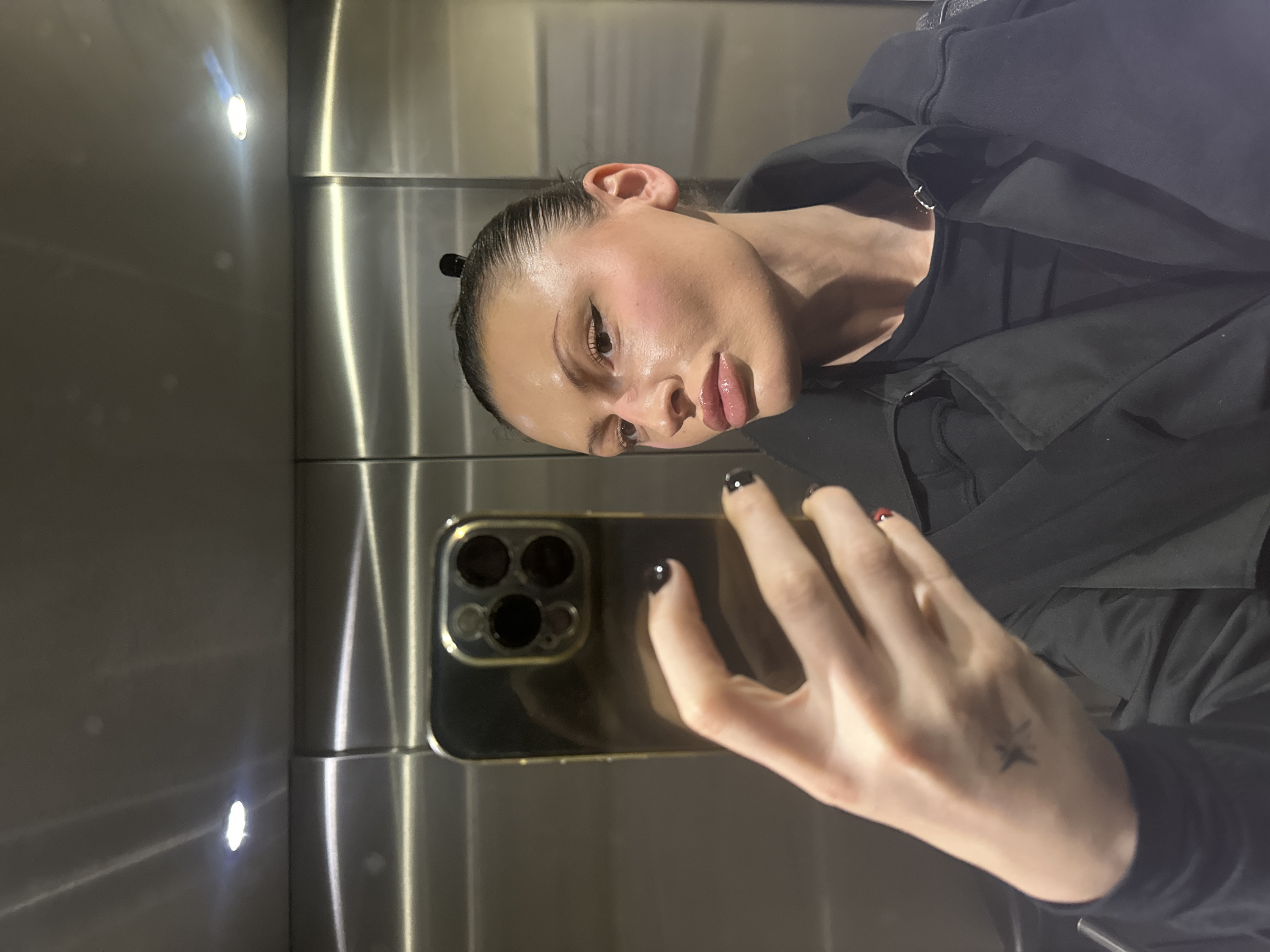 A female subject is taking a selfie within a brushed metallic elevator, located in London, United Kingdom. The elevator interior features reflective stainless steel panels with visible vertical seams and overhead circular LED lighting. The subject, positioned on the right side of the frame, is angled to her left, looking towards the screen of a gold-cased smartphone held in her left hand. The smartphone's rear exhibits a prominent triple-lens camera array. Her fingernails are painted black, with a subtle red detail visible on one finger. A small, dark, asterisk-shaped tattoo is present on the back of her left hand. Her dark hair is pulled back, revealing a small dark clip, and she displays winged eyeliner and glossy lips. She is dressed in a black collared jacket worn over a dark garment.