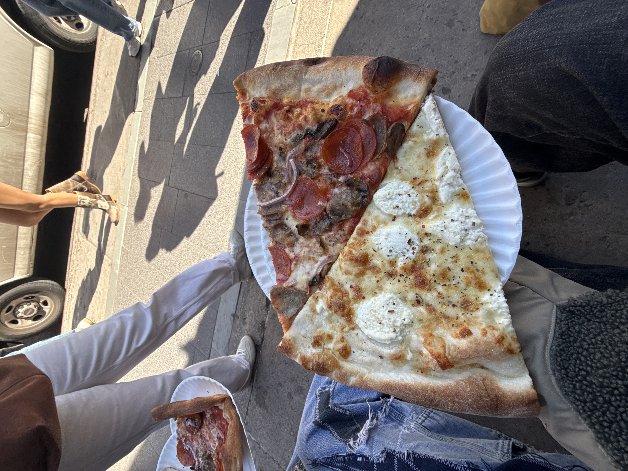 The image shows an overhead shot of two slices of pizza on a white paper plate, held between the legs of two people. One slice is topped with pepperoni, mushrooms, and onions, while the other is white pizza with ricotta-like cheese and herbs. The legs of the person on the left are clad in white pants and sneakers. They also hold a plate with a smaller slice of pizza. The person on the right is wearing jeans with a patchwork design and a beige jacket with a textured collar. The setting appears to be an outdoor location in New York City, United States of America, based on the city sidewalk and shadows that show other people standing nearby. A partial view of a car tire can be seen in the background on the left.