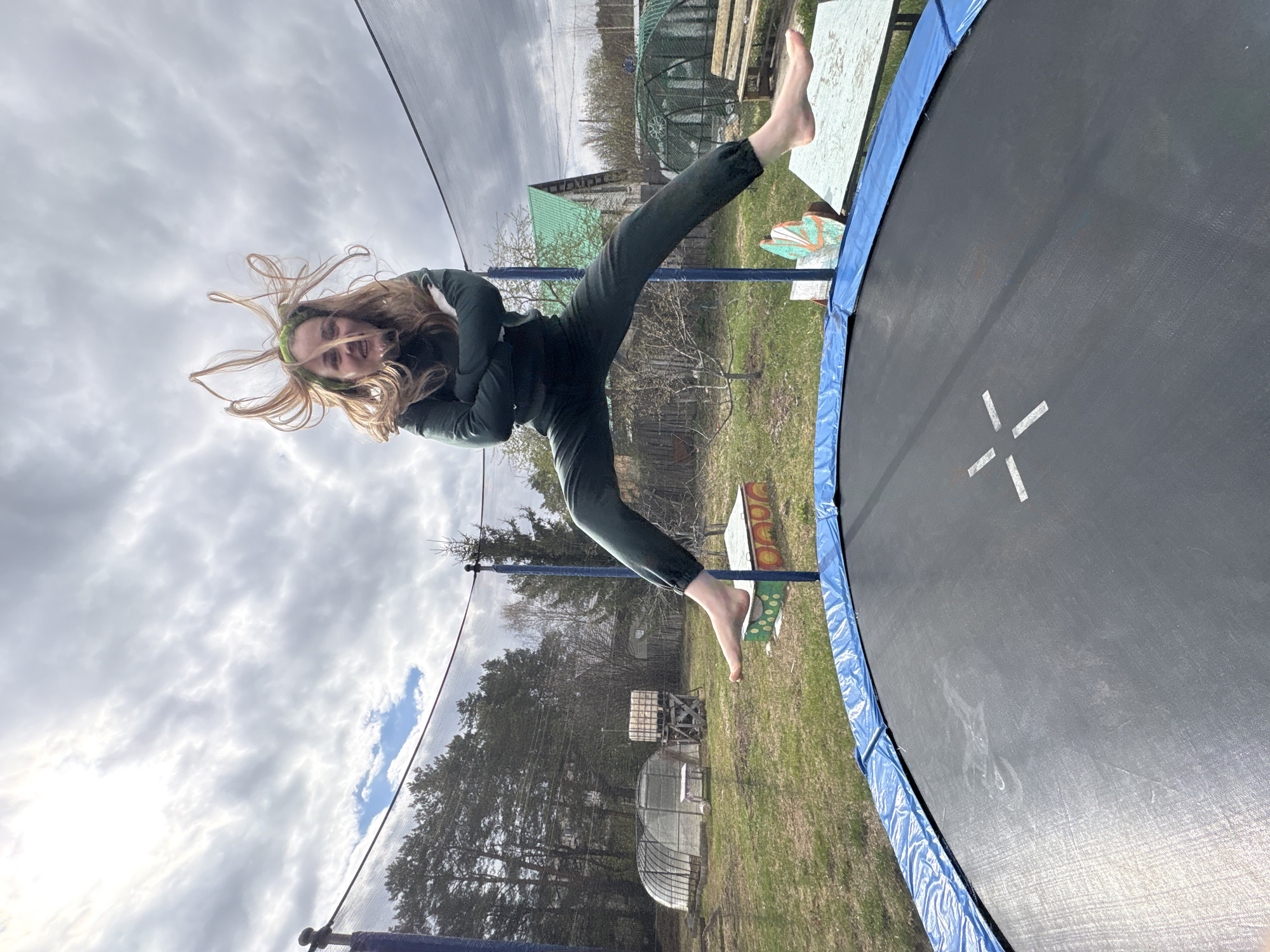 A person is captured mid-air on a trampoline. The individual is wearing dark green attire, with their arms crossed and legs extended. They are barefoot. Their long hair is dynamically spread, indicating movement. The trampoline has a blue padded edge and white markings. The surrounding environment suggests a rural or semi-rural setting, featuring a lawn, trees, and structures that might be greenhouses or outbuildings. The sky is overcast with scattered clouds. The location appears to be in Chaikovsky, Russia.