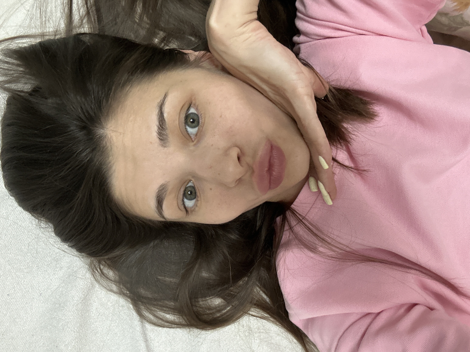 A selfie of a woman with long, dark hair, wearing a pink sweatshirt, lying on a white surface. Her left hand is resting on her face. Her eyes are light-colored. No information suggests this image was taken in Izhevsk, Russia.