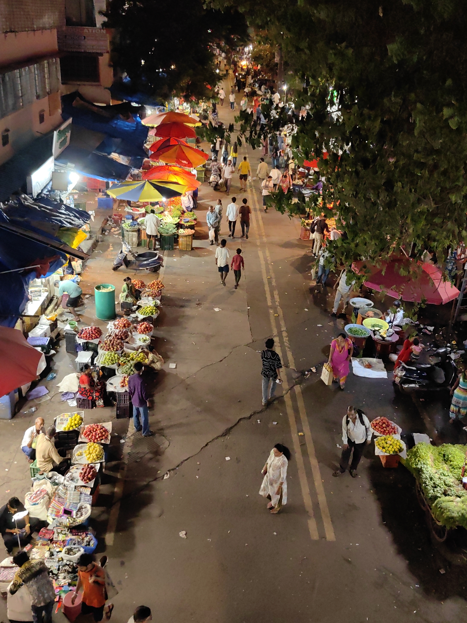 The image depicts a bustling street market in Mumbai, India, likely at night or dusk. The scene is viewed from an elevated perspective, looking down onto a street lined with vendors and shoppers. Colorful umbrellas, some red and yellow, provide shade over various stalls displaying produce and other goods.
Numerous people are present, engaged in activities such as shopping, selling, and walking. Many are dressed in traditional Indian attire. The market appears to be set up along the side of a road, with vehicles like scooters parked nearby. The ground is paved, and there are signs of litter scattered around. The buildings bordering the market are multi-story structures, some illuminated. The overall impression is one of a vibrant and active urban marketplace.