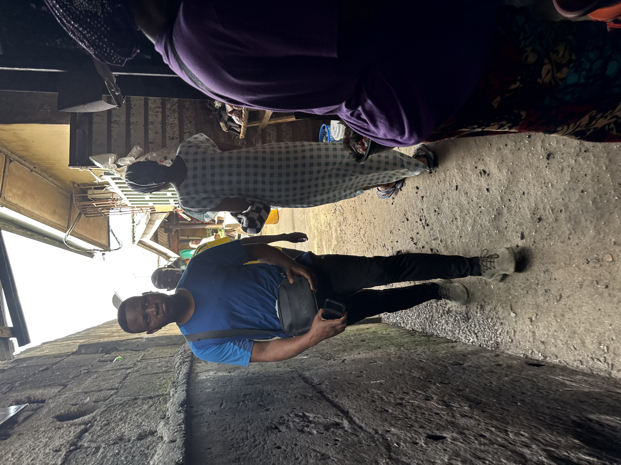 The image depicts a narrow street in what appears to be a crowded area, possibly a market or commercial zone. The man is holding a mobile phone and a bag with the initials "PO", suggesting a brand name. He is looking directly at the camera. Other people are present in the background. The street itself is paved and shows signs of wear. Overhanging structures provide partial shade. The location appears to be Lekki, Nigeria, judging from the overall environment.