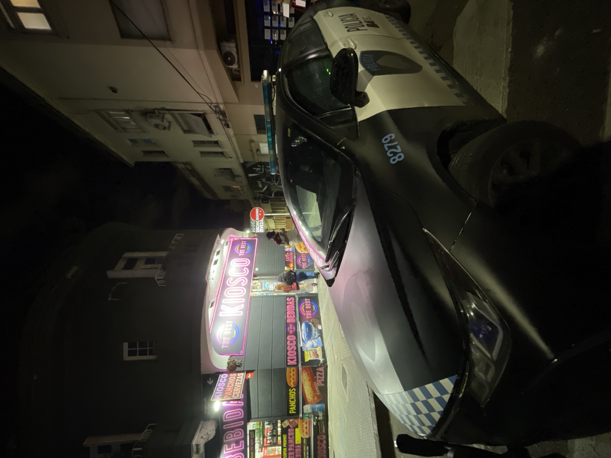 The image shows a police car parked on a street in Buenos Aires, Argentina, next to a brightly lit kiosk called "The Best Kiosco." The kiosk has signs advertising various food and drinks, including "Bebida" (drinks), "Panchos" (hot dogs), "Cervezas" (beers), and "Pizza." People are standing in front of the kiosk. The police car has the number "8279" visible on its side. Buildings are visible in the background. The photo was taken at night.