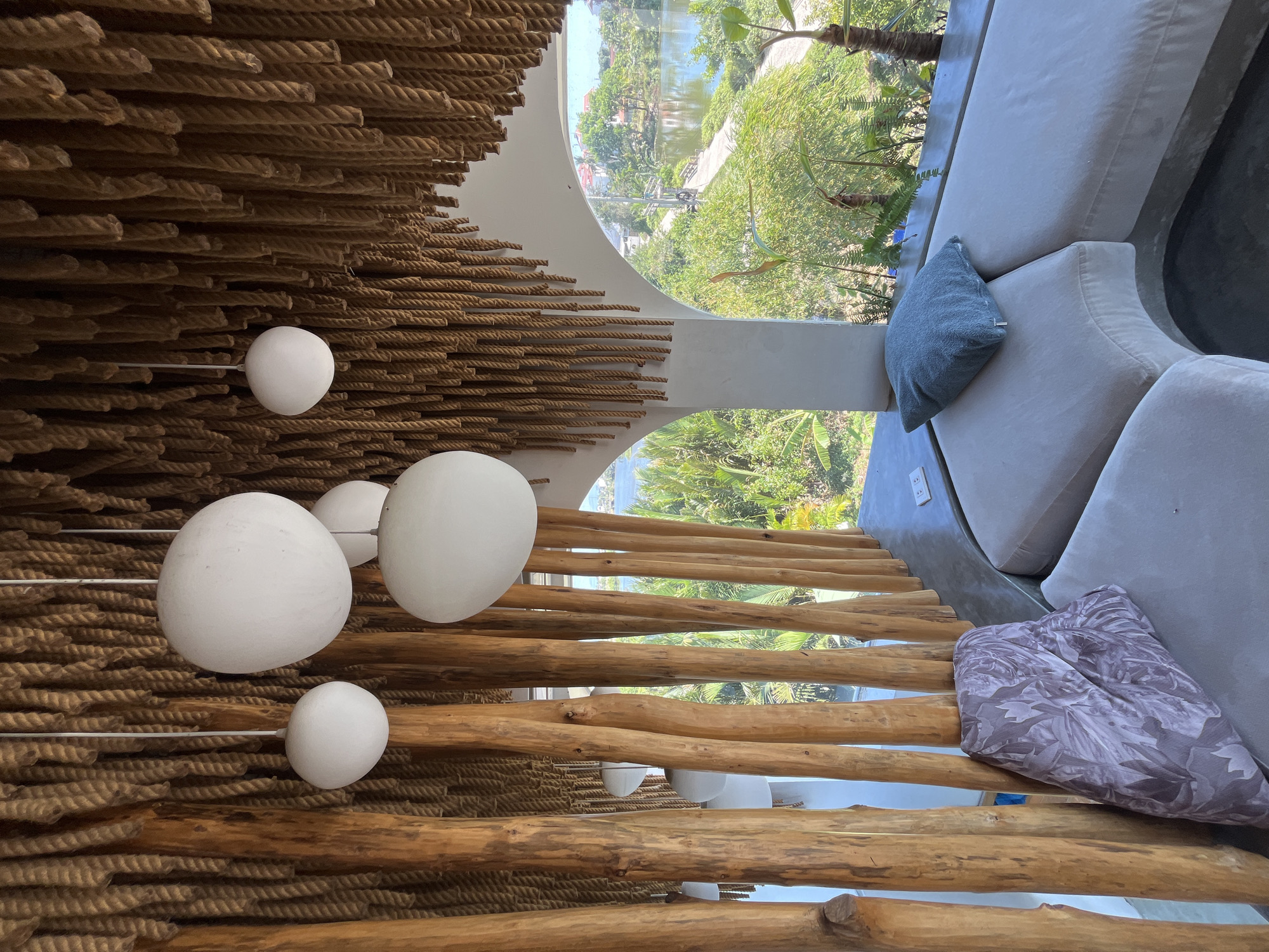 The image depicts an inviting, modern interior space with a strong connection to the outdoors, characteristic of a resort or luxury residence, likely in Đà Nẵng, Vietnam. The perspective is rotated 90 degrees clockwise, showing the scene as if viewed from above and to the side.
The left and upper portions of the frame are dominated by a unique architectural or decorative element. This features rows of thick, natural brown rope arranged horizontally, interspersed with horizontal wooden logs. From this textured surface, several white spherical light fixtures of varying sizes are suspended by thin rods, adding a soft, contemporary glow to the rustic material.
Towards the right and lower sections of the image, a large, curved window or open arch provides a clear view of a lush outdoor environment. Beyond the glass, vibrant green foliage and trees are prominent, with a body of water, possibly a river or pond, visible in the distance. The outdoor setting suggests a tropical or subtropical climate.
Inside, a comfortable, light-colored, curved sofa is positioned to enjoy the view. Two throw pillows adorn the sofa: one is a plain light blue or teal, and the other features a greyish-purple floral or leafy pattern. The flooring or supporting structures around the seating appear to be a smooth, light grey material.
The scene is bathed in bright daylight, indicating a sunny day. No people are visible, suggesting a peaceful and serene atmosphere designed for relaxation. There is no visible text in the image.