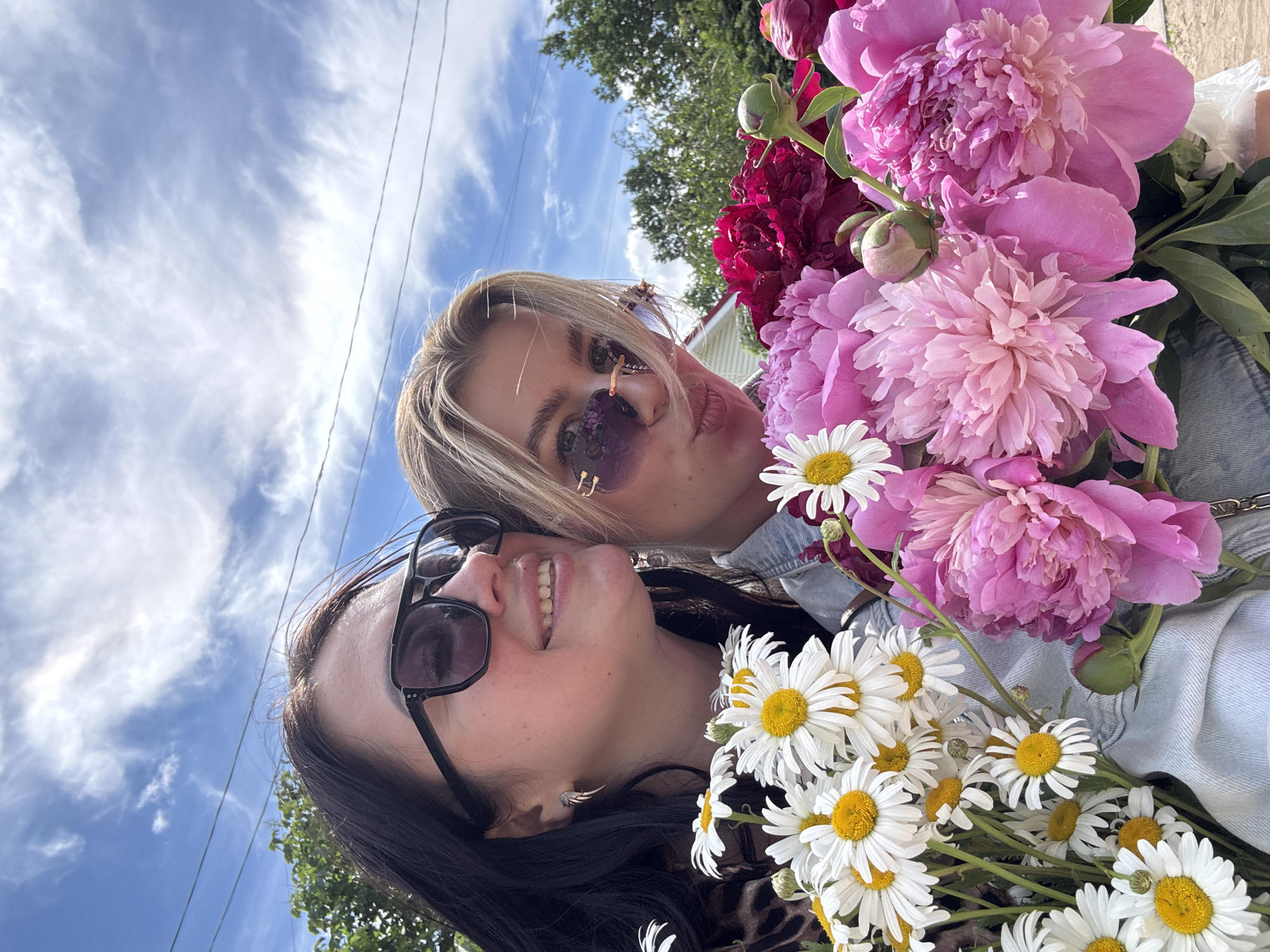This image is a brightly lit, rotated selfie featuring two women holding an abundance of flowers outdoors on a sunny day. The original photo is oriented so that the sky is on the left and the subjects are on the right, but it depicts two individuals holding flowers against a sky background.
In the foreground, a woman with dark hair and dark sunglasses is smiling broadly towards the camera, her teeth visible. She is wearing a dark top, possibly with a leopard print, and a denim jacket. A large cluster of white daisies with yellow centers is prominently displayed in front of her.
Behind her, positioned slightly above and to the right, is another woman with blonde hair and purple-tinted sunglasses. She is making a playful kissing face towards the camera. She also appears to be wearing a denim jacket. She is surrounded by a large, lush bouquet of pink and dark red peonies, with some unopened peony buds visible.
The background reveals a clear blue sky with scattered white, wispy clouds and a few utility wires stretching across the frame. Green tree foliage is visible at the top and bottom edges (when upright), suggesting an outdoor setting like a park or residential area. The bright lighting indicates it is daytime. Both women appear happy and are captured in a moment of friendly interaction, adorned with colorful flowers. There is no visible text in the image.