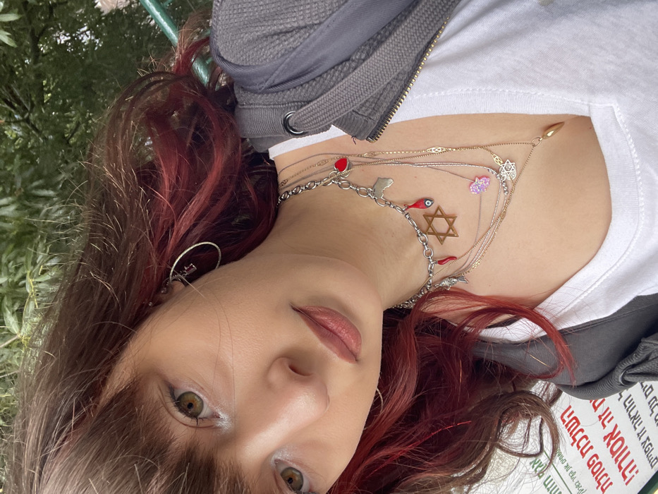 A close-up selfie depicts a young female individual outdoors in Raanana, Israel. The subject has brown hair with prominent reddish-magenta streaks and bangs, hazel eyes, and wears lip color. She is dressed in a white v-neck t-shirt beneath a partially unzipped dark grey hooded jacket. Adorning her neck are multiple layered necklaces featuring various charms, including a silver-colored chain with a red heart, an outline of the African continent, a red chili pepper, a small fish, a wooden Star of David, and an eye charm. Additional delicate chains carry a pink Hamsa hand, a white Star of David, and a gold tear-drop pendant. A silver-colored hoop earring with a small dark charm is visible on her right ear. The background consists of lush green foliage in the upper section and a white sign with Hebrew text on the lower left, partially visible, reading "השלכת פסולת זה אסור!!" (Disposing of waste is forbidden!!). The scene is illuminated by natural daylight.