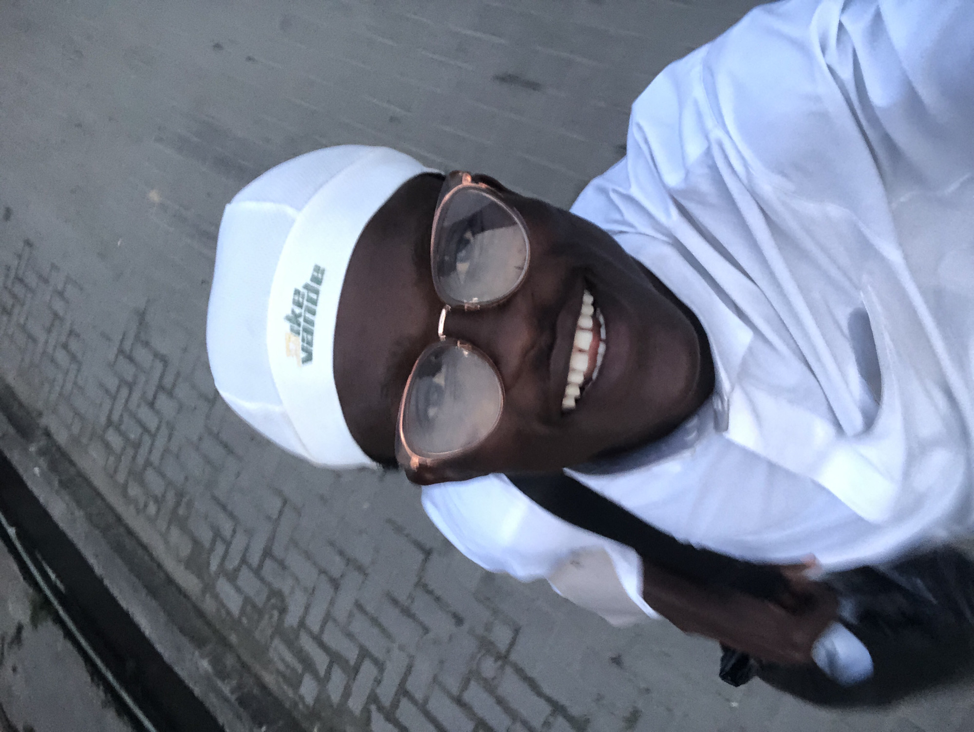 A person wearing a white cap with the logo "Nike Vande" and a white shirt takes a selfie outdoors on a paved surface. The person is smiling widely, revealing their teeth, and wearing rose-gold framed glasses. Part of the person's dark hair is visible under the cap. They are carrying a black bag over their left shoulder. The background consists of a gray concrete pavement with a pattern of interlocking rectangular bricks, and a dark curb with a thin dark line along its edge, suggesting an outdoor urban environment, possibly a walkway or road in Ado, Nigeria. The lighting indicates it may be late afternoon or early evening.