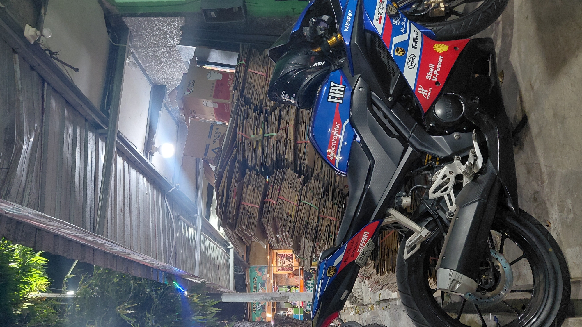 In Bantarsoka, Indonesia, a blue and black motorcycle is parked outdoors under a corrugated metal awning. The motorcycle has several stickers and logos, including "Santander," "FIAT," "UPS," "Ferrari," "Shell V-Power," and "Kaspersky." A black helmet rests on the seat of the motorcycle. Behind the motorcycle, a large stack of flattened cardboard boxes is piled up. In the background, a small food stall with signage is visible, and a person is standing inside. Trees with green leaves are also present, and the lighting suggests it is evening or nighttime, with artificial lights illuminating the scene. The ground appears to be paved or concrete.