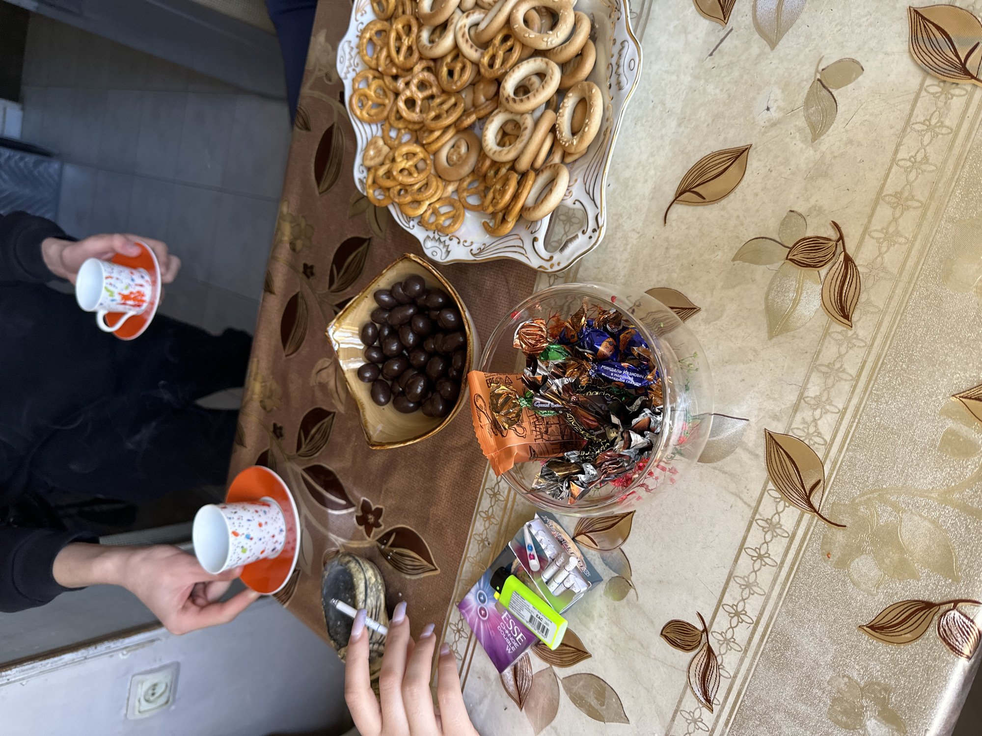 The scene depicts a tabletop setting, likely in a home, with the following elements:
**People:** Two individuals are partially visible. One is holding a cup and saucer. Another person is holding a lit cigarette.
**Objects:**
* A tray filled with bagels and pretzels.
* A small bowl of chocolate-covered items.
* A glass bowl filled with candies.
* A packet of cigarettes ("ESSE" brand is visible).
* An ashtray with a cigarette.
* Two cups with saucers.
* The tabletop is covered with a decorative tablecloth with floral and leaf patterns.
**Actions:**
* Someone is holding a lit cigarette, possibly smoking.
* Another person is holding a cup and saucer.
**Environment:**
* The setting is indoors. The tablecloth suggests a domestic environment.
The specific location in Yerevan, Armenia, cannot be determined with only the provided information.