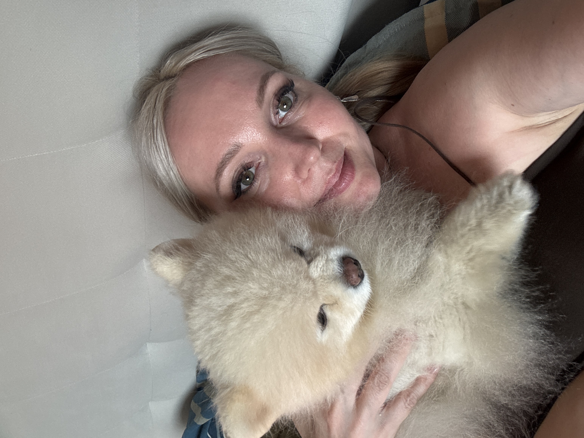 A woman with blonde hair and green eyes, wearing makeup, is holding a light-colored Pomeranian dog. The woman is looking at the camera, and the dog is nestled in her arms. They appear to be on a couch or bed, as suggested by the soft, textured background and a glimpse of a patterned blanket. The lighting suggests an indoor setting, possibly a home. The image was taken in Komsomolsk-on-Amur, Russia.