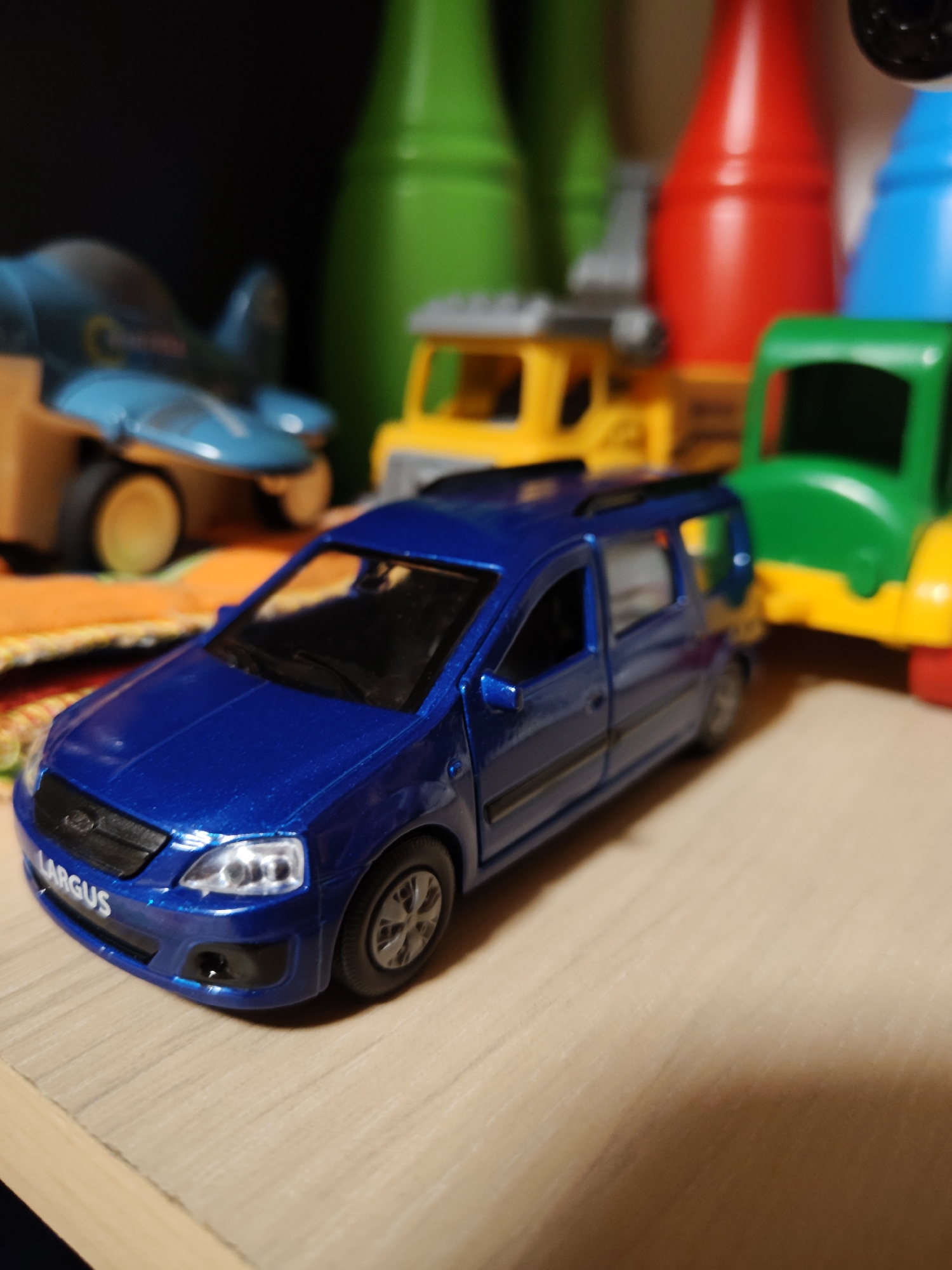 In the city of Фестивальное Снт, Russia, a close-up, eye-level shot captures a collection of toy vehicles arranged on a wooden surface. The primary focus is a metallic blue toy car, specifically a Lada Largus, positioned in the foreground. The car's headlights are illuminated, and the "LARGUS" text is visible on its front bumper. Behind the blue car, to the left, is a partially visible blue toy airplane with yellow accents and visible wheels. Further to the right, a bright yellow toy truck is visible, followed by a green toy truck with a yellow chassis. In the blurred background, a red, green, and blue cylindrical object, possibly representing bottles or similar toys, are present. The overall scene suggests a playful or display setting for toy vehicles.