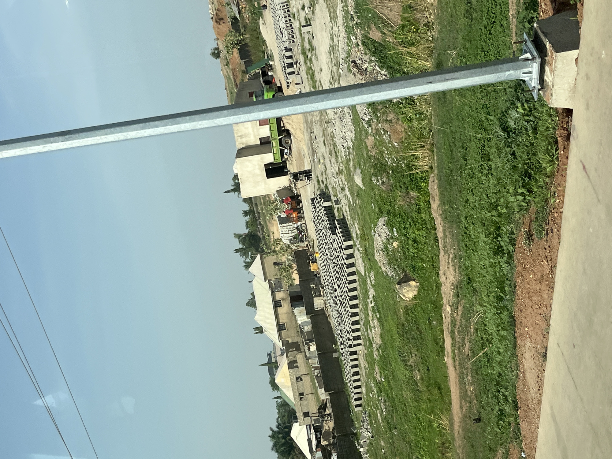 This image captures an outdoor scene at a construction or development site in Uke, Nigeria, under a clear, bright sky. The photograph is taken from a slightly elevated viewpoint, looking down upon the area. A prominent, silver metallic pole, likely a street light or utility pole, runs diagonally from the lower right foreground towards the upper left, partially obscuring the view.
In the midground and background, several unfinished buildings constructed from concrete blocks are visible. Some of these structures have rudimentary roofs, suggesting residential or commercial development. Adjacent to these buildings and spread across the site are large, neat stacks of grey concrete blocks, indicating active construction. Two green trucks are present: one is partially hidden behind a building on the left, and another is visible further back within the site.
The ground is a mix of bare earth, dirt paths, and patches of green grass and weeds. There are also areas with scattered debris and rubble typical of a construction zone. The weather appears sunny with a light blue sky, indicating daytime, possibly late morning or early afternoon, given the lack of harsh shadows. No people are visible within the frame, and there is no discernible text. The scene conveys ongoing development and building activity.