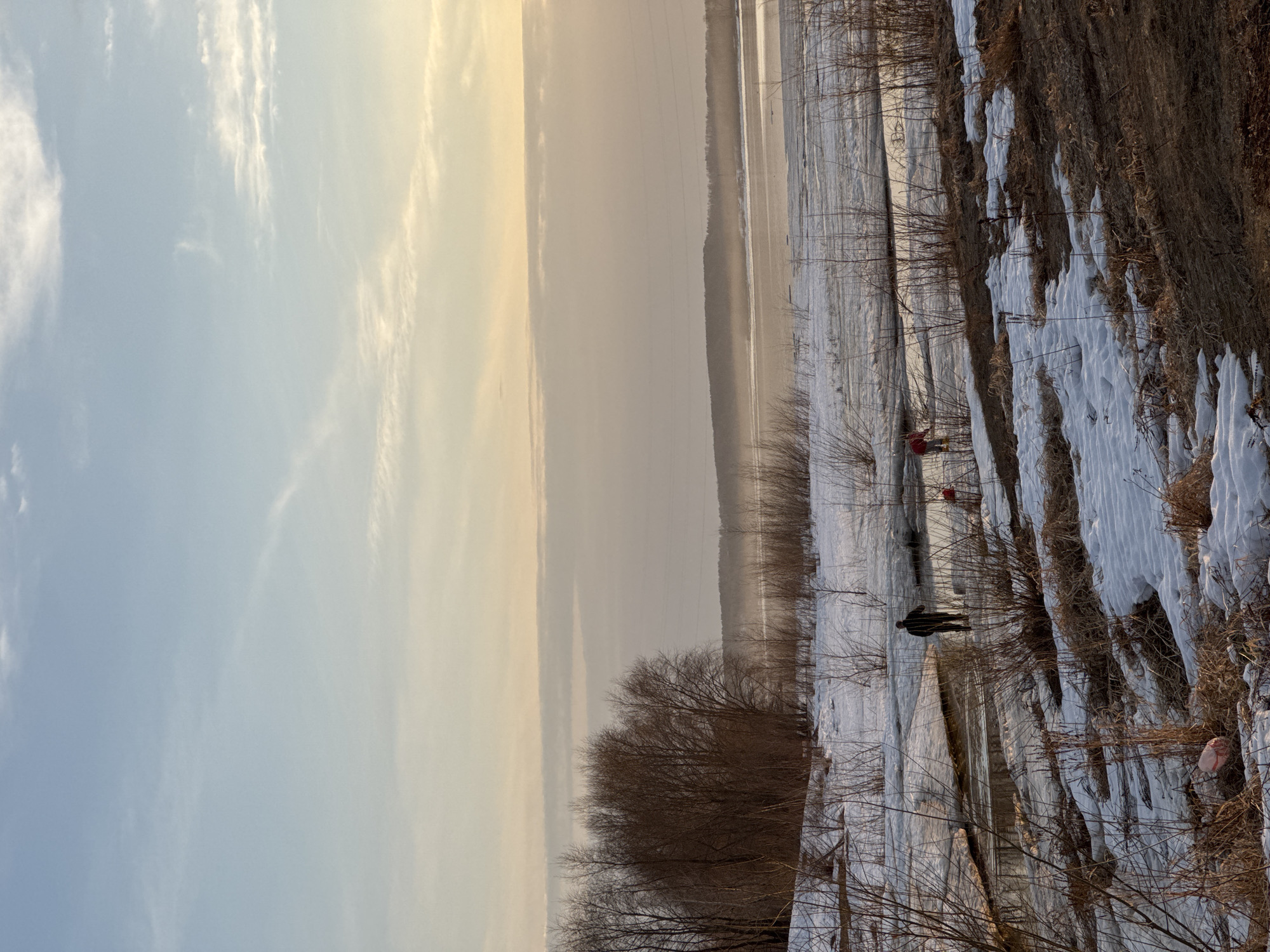 The image depicts a landscape in Chaikovsky, Russia, during what appears to be early spring or late winter, with remnants of snow covering the ground. The sky is pale blue and yellow. A body of water, possibly a partially frozen river or lake, stretches across the middle ground. Bare trees and brush line the edge of the water. On the right side, snow and dry grass cover the bank. Two figures are visible: one dressed in black stands near the water's edge, and another in red is further away. The overall scene suggests a cold, tranquil environment.