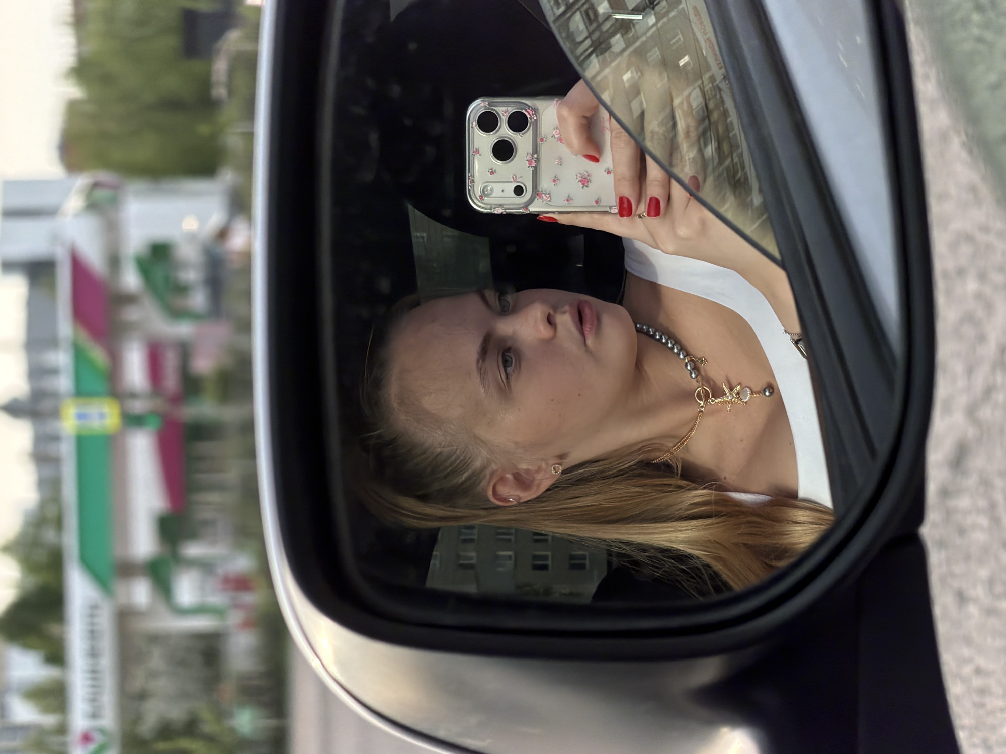 A fair-skinned female takes a selfie in a vehicle's side mirror, featuring an iPhone with a floral case and red nail polish. She is wearing a white top and a pearl necklace with a golden starfish charm. The reflection in the mirror shows a blurred urban backdrop, specifically a building and a gas station identifiable as a "Лукойл" location in Izhevsk, Russia. The photo orientation is rotated sideways.