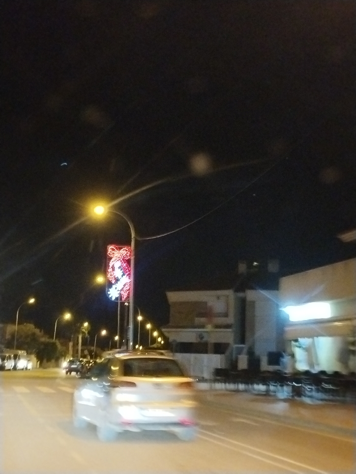 A blurred image captured at night shows a light-colored car in motion on a city street. Street lights illuminate the scene, with one pole featuring a decorative light display. Buildings are visible on the right side of the road, with what seems to be a cafe or restaurant with outdoor seating. Other cars are seen in the distance, creating a sense of urban activity. The dark sky contrasts with the artificial lights of the cityscape.