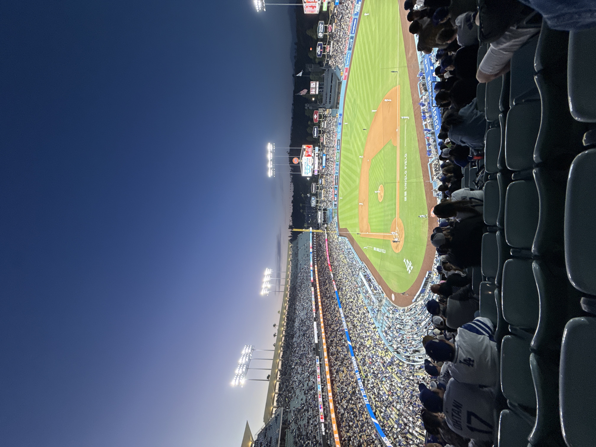 This image shows a baseball game at Dodger Stadium in Los Angeles, California. The stadium is full of spectators. The baseball field is visible with various markings and the word "UNIQLO FIELD". The sky is a dark blue. The view is from a high vantage point, likely from seats in the stadium, looking down onto the field and surrounding seating areas. Spectators are wearing various clothing including Dodger jerseys, one with the name "OHTANI 17" visible. Empty seats are in the foreground of the image. Stadium lights illuminate the field. The game is taking place during the evening.