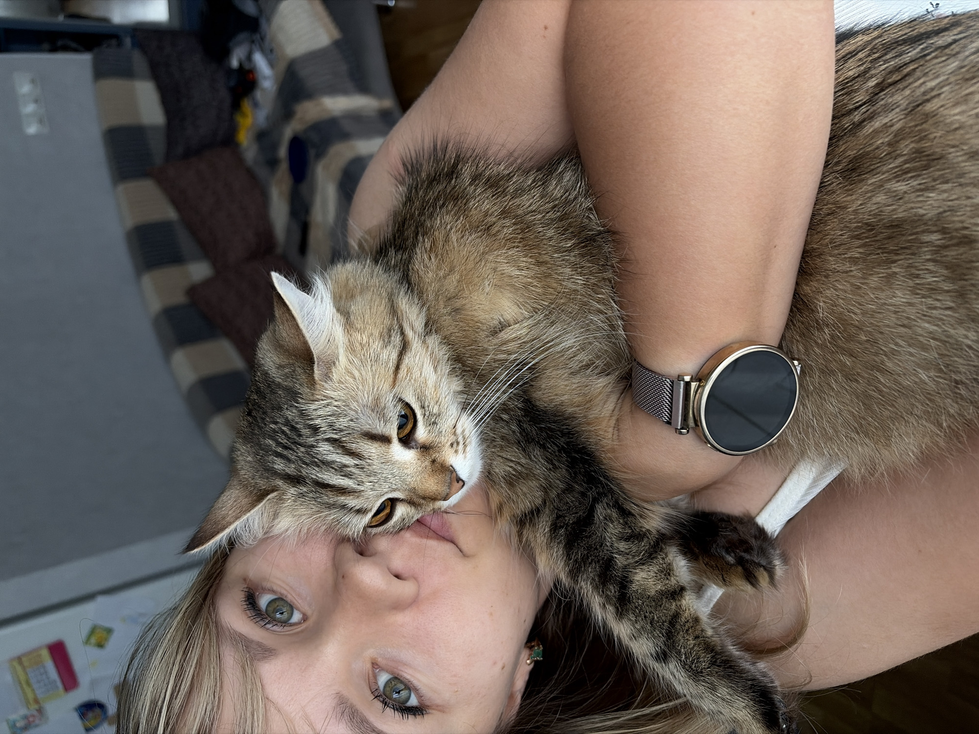 A light-skinned woman with blonde hair and green eyes is holding a brown tabby cat close to her face. She is wearing a smart watch on her left wrist. The cat is positioned with its head near her face, almost touching her lips. The background shows a couch with a patterned blanket. The location is Chaikovsky, Russia.