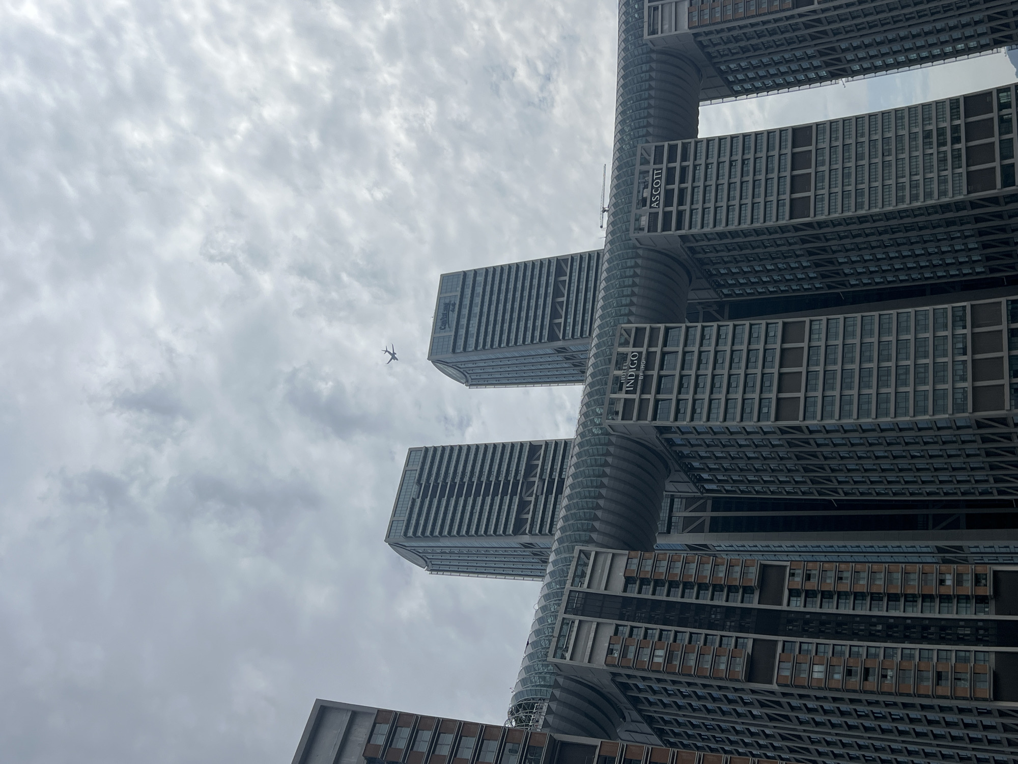 This is a vertical, low-angle shot of the Raffles City Chongqing complex in Yuzhong District, China, on an overcast day. The complex consists of eight towering skyscrapers connected by a horizontal glass-enclosed walkway named "The Crystal". In the sky, a small airplane is visible in flight, appearing as a silhouette against the cloudy backdrop. The architecture of the skyscrapers is modern and angular, with numerous windows. Signs for "HOTEL INDIGO" and "ASCOTT" are visible on the buildings. The overall environment appears to be urban, with a focus on the monumental scale of the architecture. There are no people or visible interactions depicted.