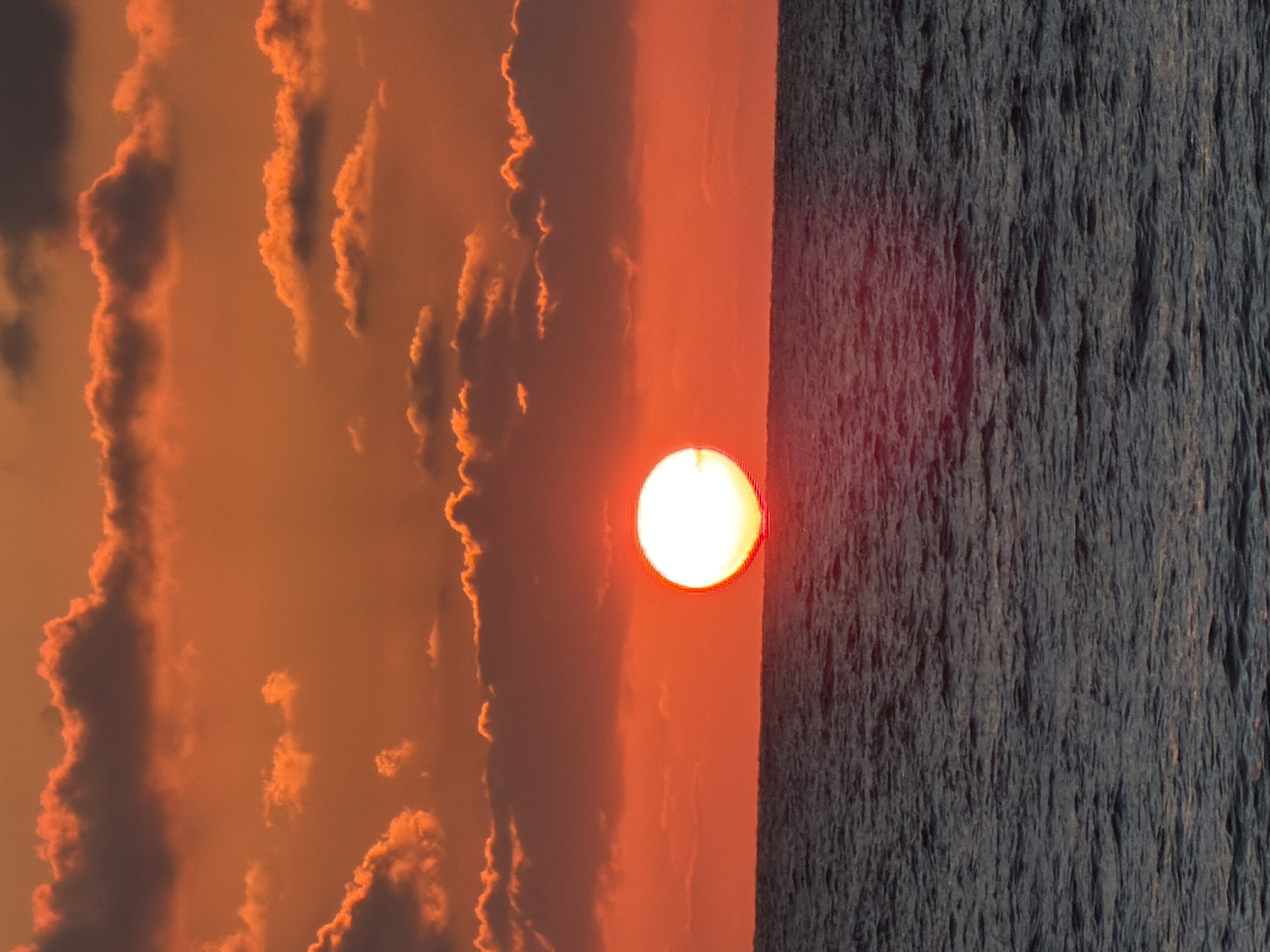 The image depicts a sunset, likely over the Mediterranean Sea, near Tel Aviv, Israel. The sun is a bright, circular orb positioned in the center of the frame. To the left of the sun are dense, orange-tinged clouds. To the right of the sun is a body of dark, choppy water. The image is rotated 90 degrees counterclockwise.